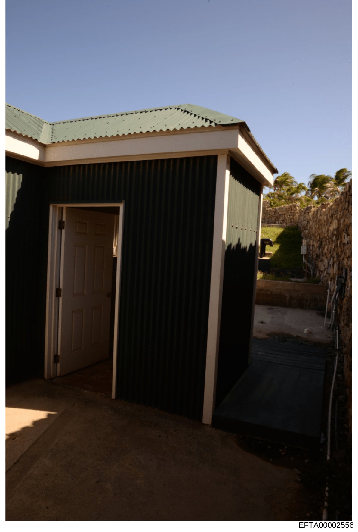 This is a photograph of a small, single-story outbuilding with corrugated metal siding and a metal roof. The structure appears to be used for storage or utility purposes, featuring a brown wooden door and situated in what appears to be a rural or sem