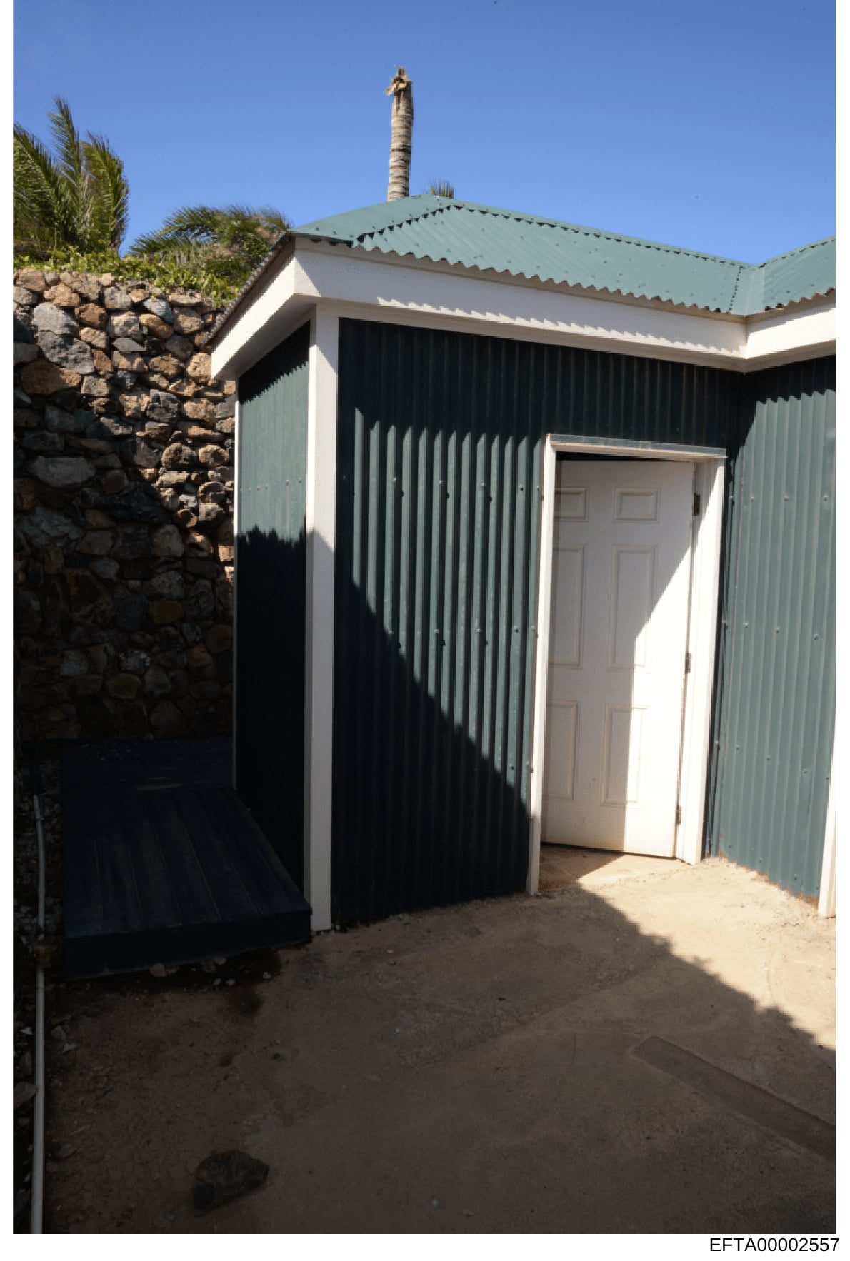 A color photograph of a small residential outbuilding or guest house featuring corrugated metal siding painted green and white, with a pitched roof and chimney. The structure appears to be located on a property with stone walls and tropical vegetatio