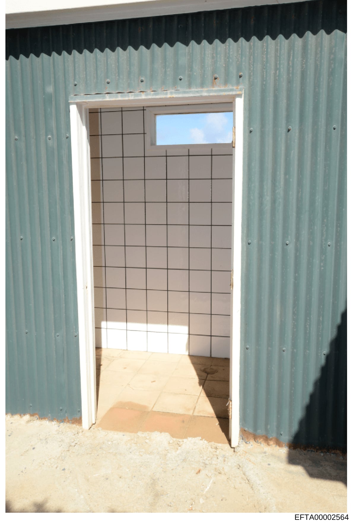 This is a photograph showing the interior of a corrugated metal building or structure, taken through an open doorway. The image reveals a room with beige/tan tiled walls in a grid pattern, a concrete floor with sand/dirt visible at the entrance, and