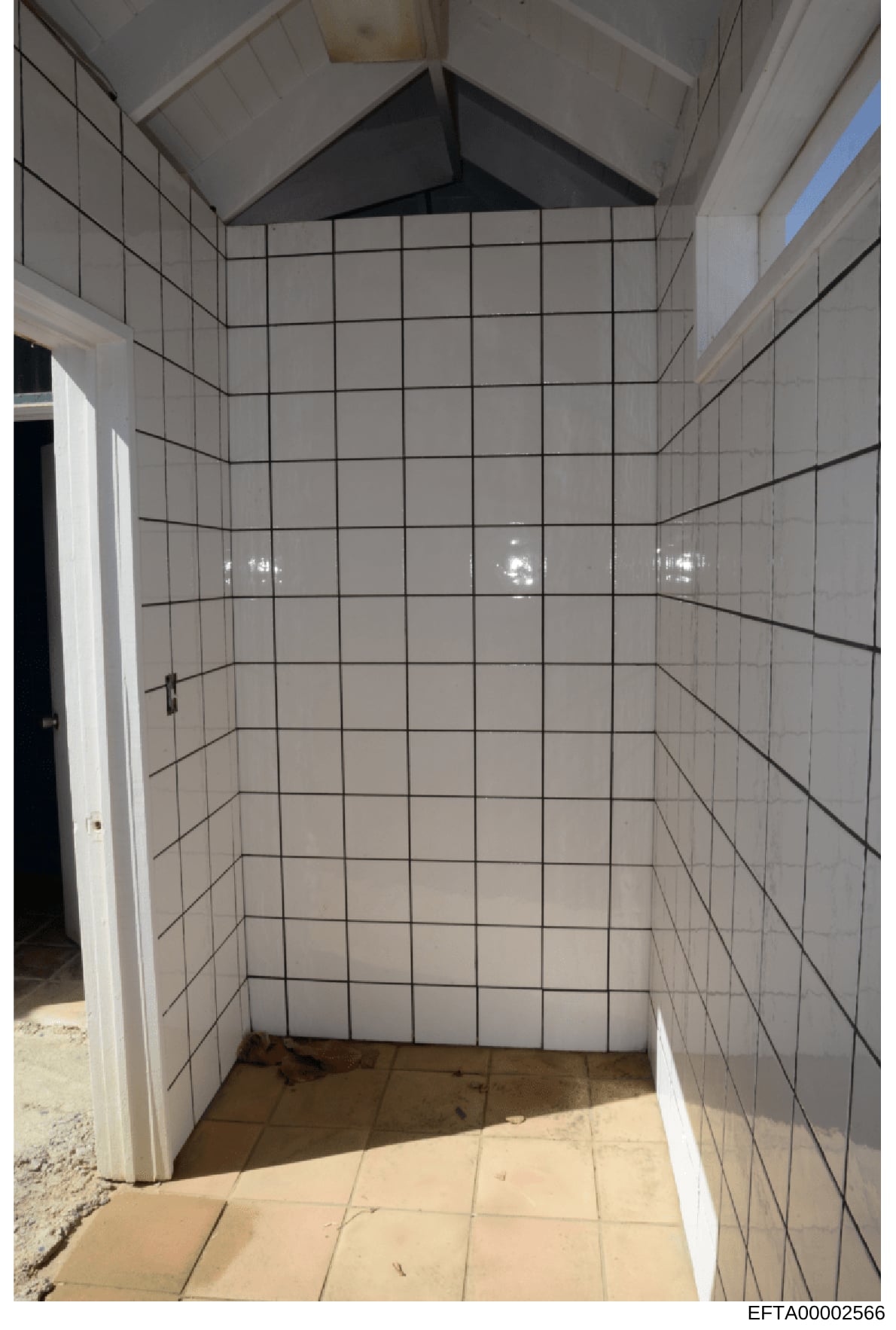 This photograph depicts an interior bathroom room with white tile walls and a vaulted ceiling. The room appears to be under construction or in a state of renovation, with minimal fixtures visible and exposed construction materials on the floor. The B