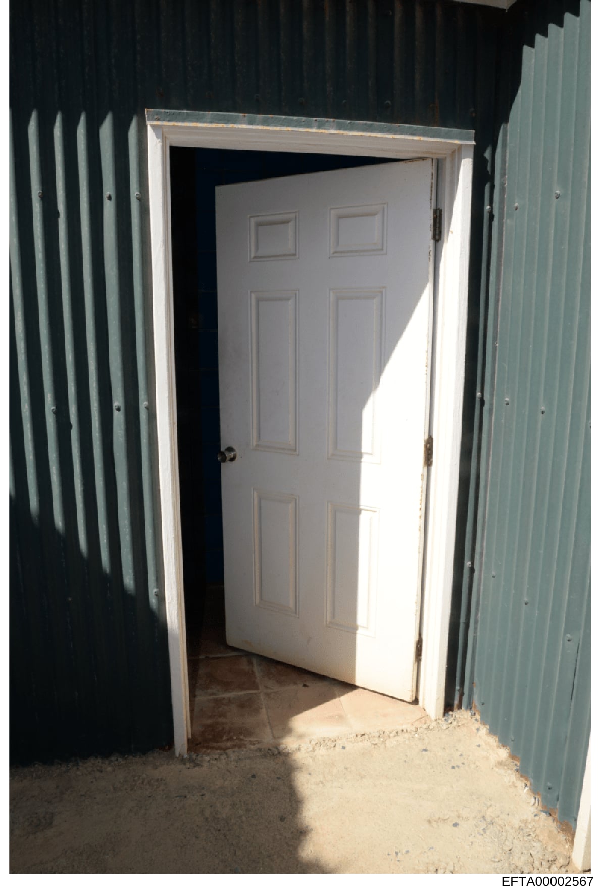 This is a photograph showing an open white six-panel interior door within a metal-framed building structure. The door appears to be standard residential or light commercial grade, with corrugated metal siding visible on both sides of the frame. The i