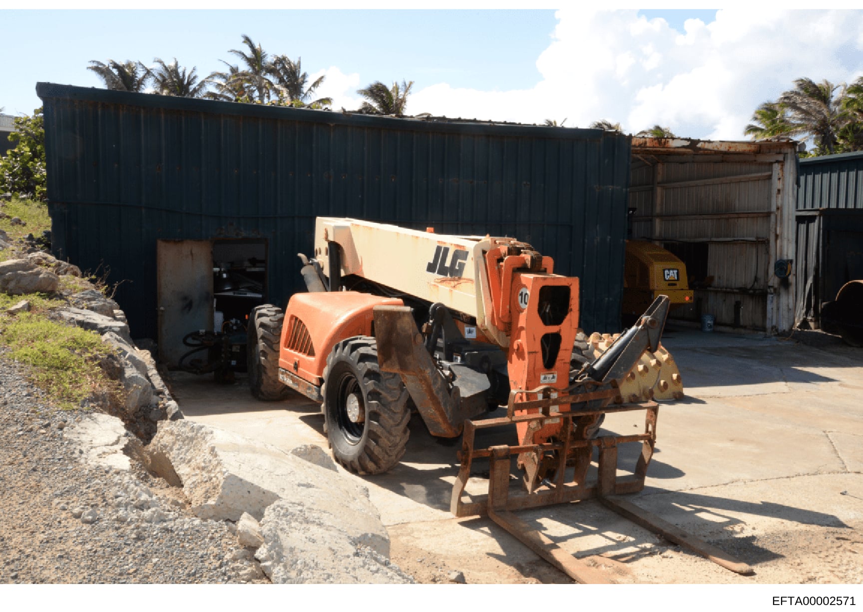 This photograph shows a property or facility containing heavy construction and industrial equipment, including a JLG telehandler, a CAT skid loader, and an orange loader vehicle, situated in front of dark metal storage buildings with corrugated metal