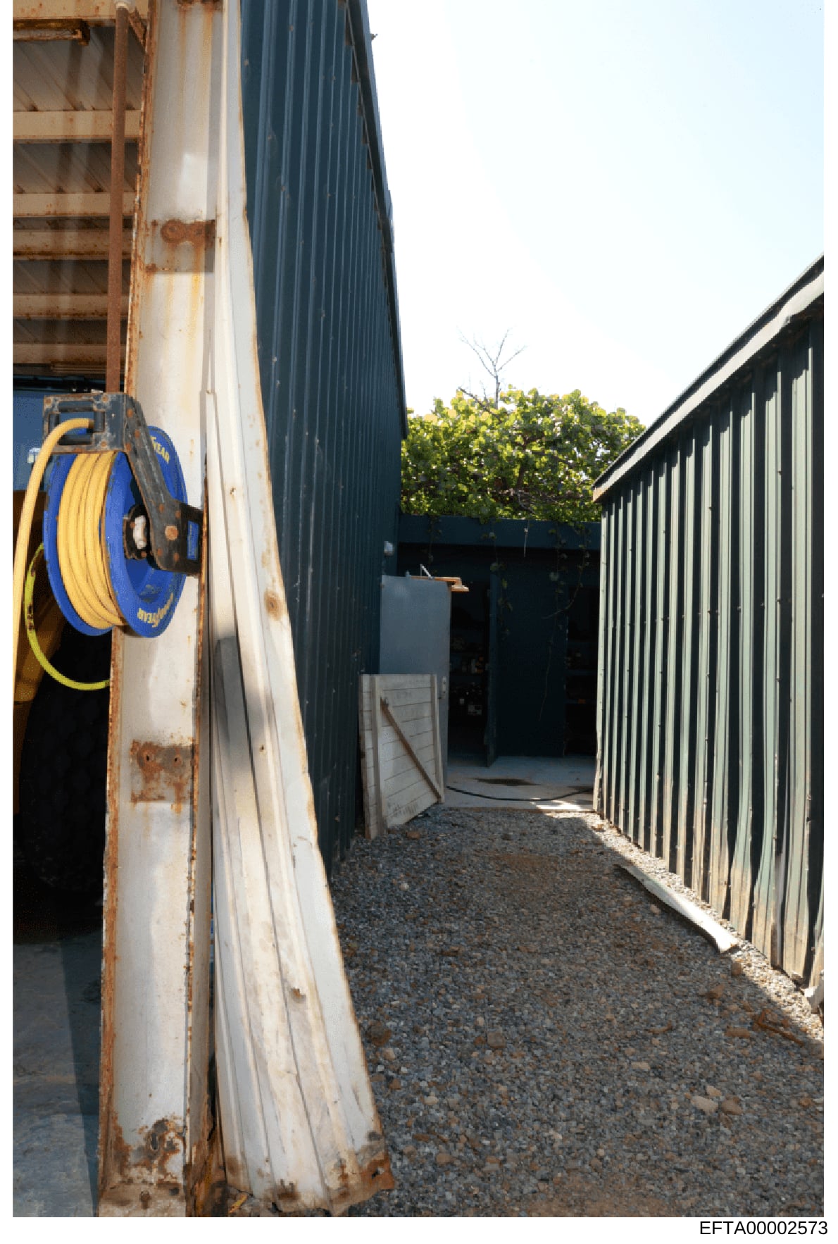 This photograph shows the exterior of an industrial or warehouse building featuring corrugated metal structures, a blue and yellow hydraulic hose reel mounted on a weathered white panel, gravel flooring, and metal fencing. The image appears to be doc