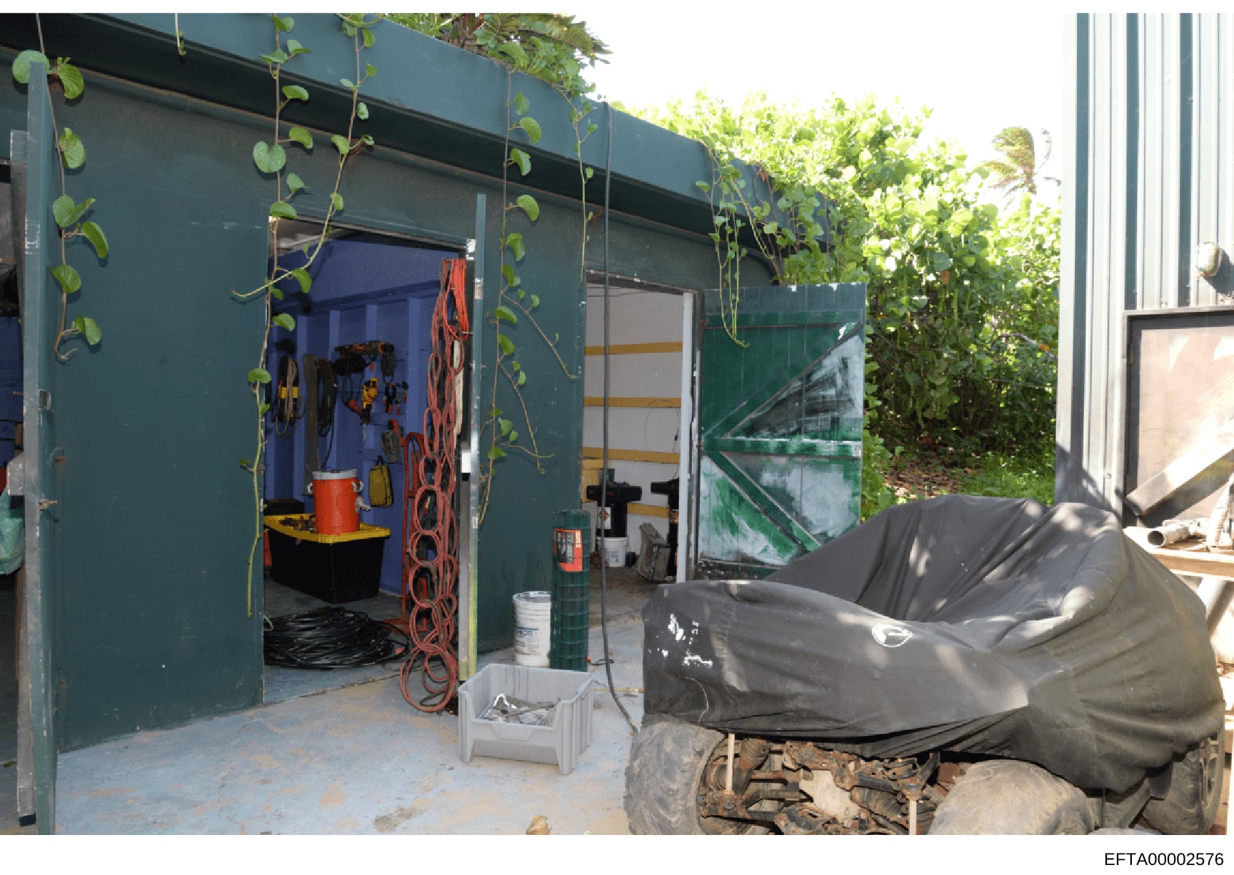 This is a photograph of an outdoor utility building or shed with a teal/dark green exterior, featuring climbing vines and vegetation. The structure contains various equipment including coiled red hoses, storage containers, buckets, and maintenance su