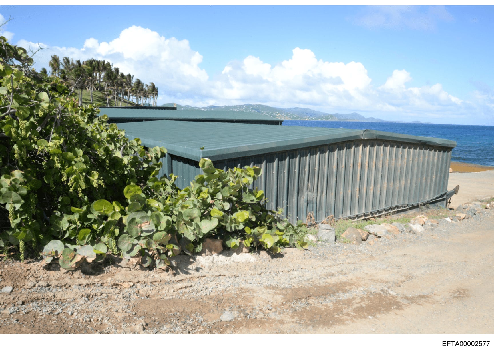Photograph of a modern metal-clad structure located on a beach, featuring a corrugated metal exterior with a sloped roof. The building overlooks a scenic coastal area with mountains visible in the distance and tropical vegetation (palm trees) in the 