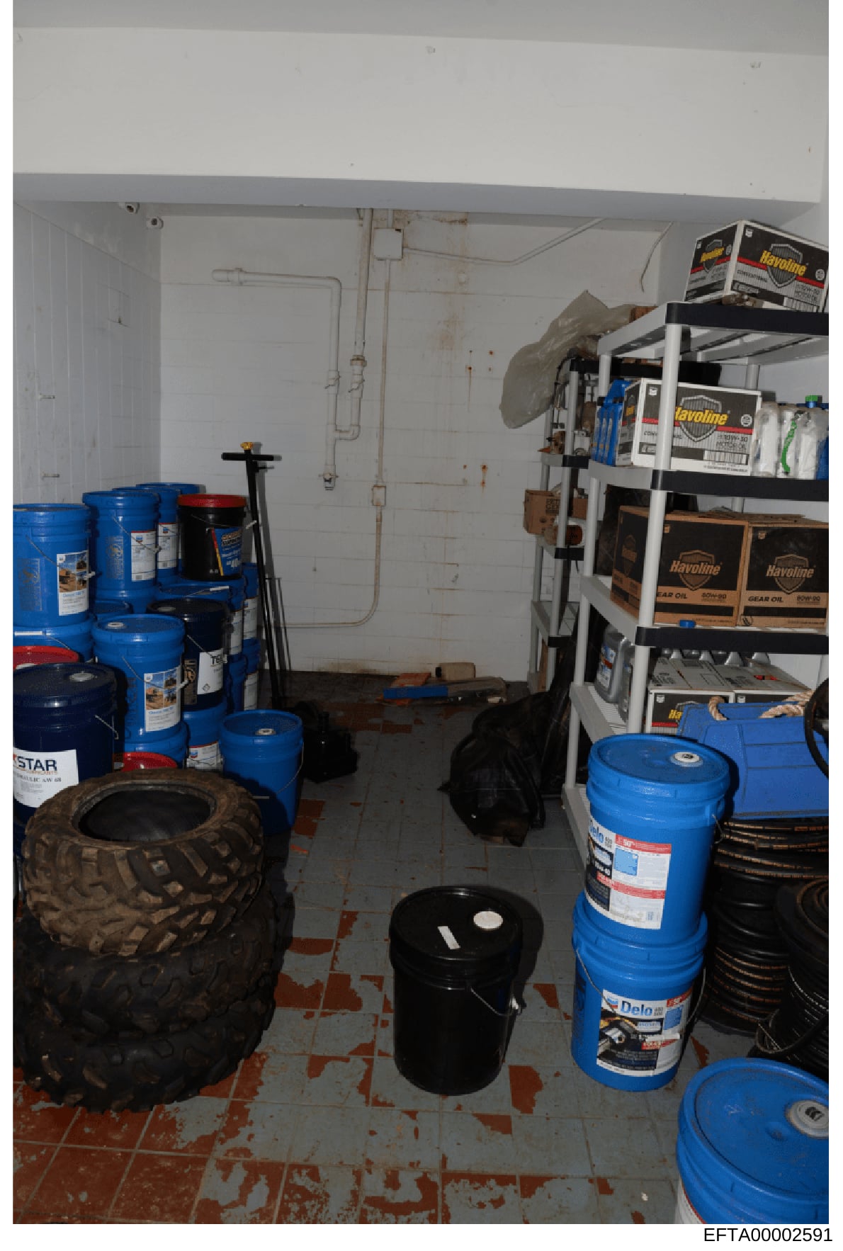 This is a photograph of a storage or utility room containing multiple large industrial drums (blue and black), automotive fluids, oils, shelving units with product boxes, a tire, and various maintenance supplies. The room shows signs of age with stai