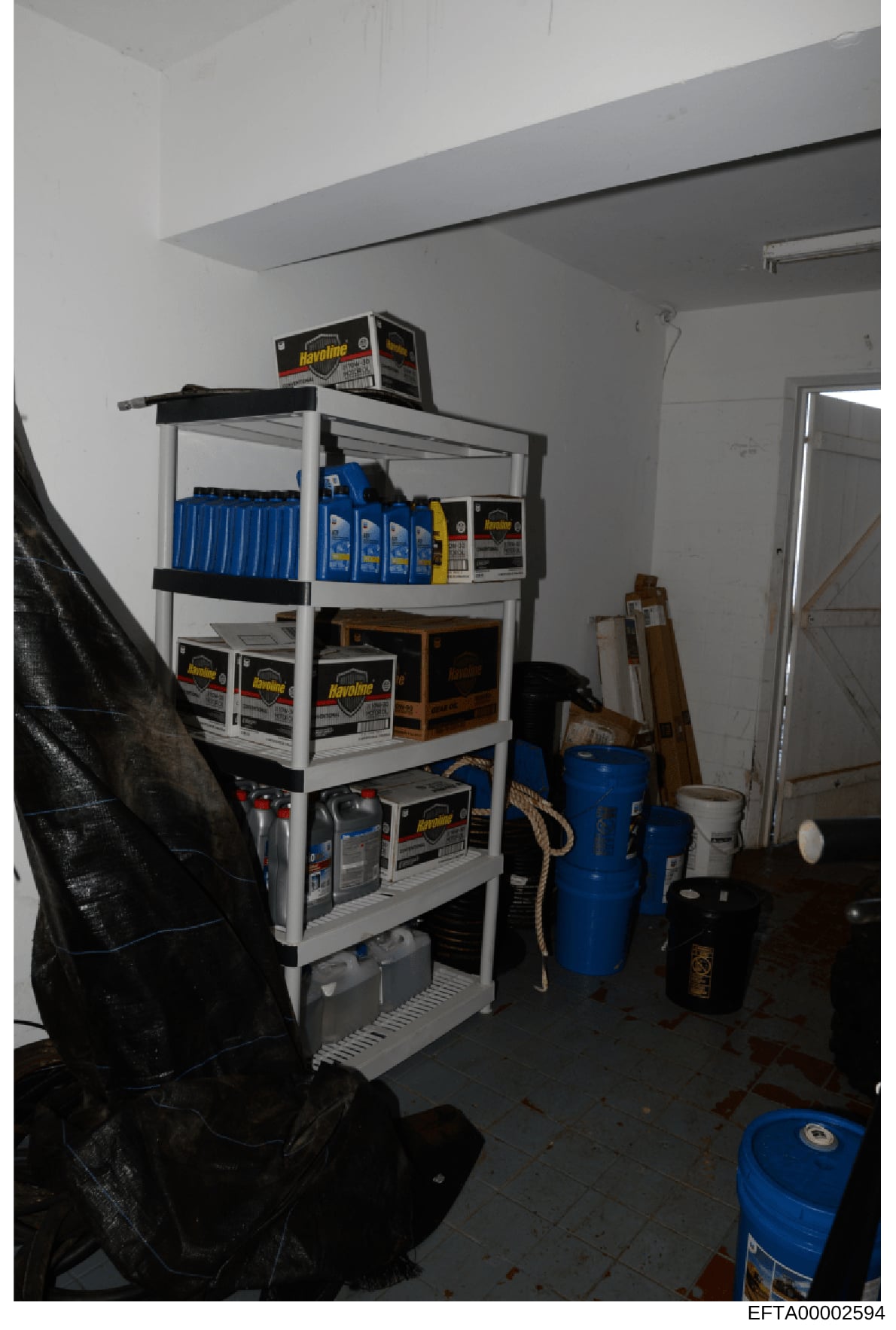 This is a photograph of a storage or utility room containing shelving units stocked with Havoline motor oil products, cleaning supplies, blue containers, and various maintenance equipment. The image appears to be evidence documentation from the Epste