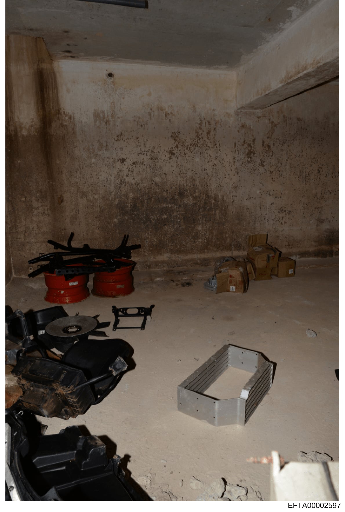 This is an evidence photograph showing an interior room or basement space containing various items including red containers, cardboard boxes, black equipment/machinery parts, and metal fixtures. The room shows signs of significant moisture damage and