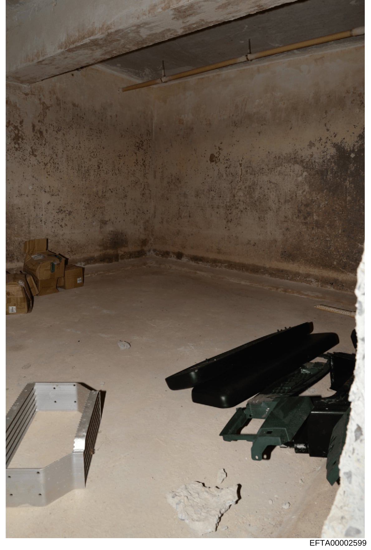 This is an evidence photograph showing an unfinished interior room with deteriorated concrete walls and floor. The space contains various items including cardboard boxes, black objects (possibly furniture or equipment), and metal/mechanical component