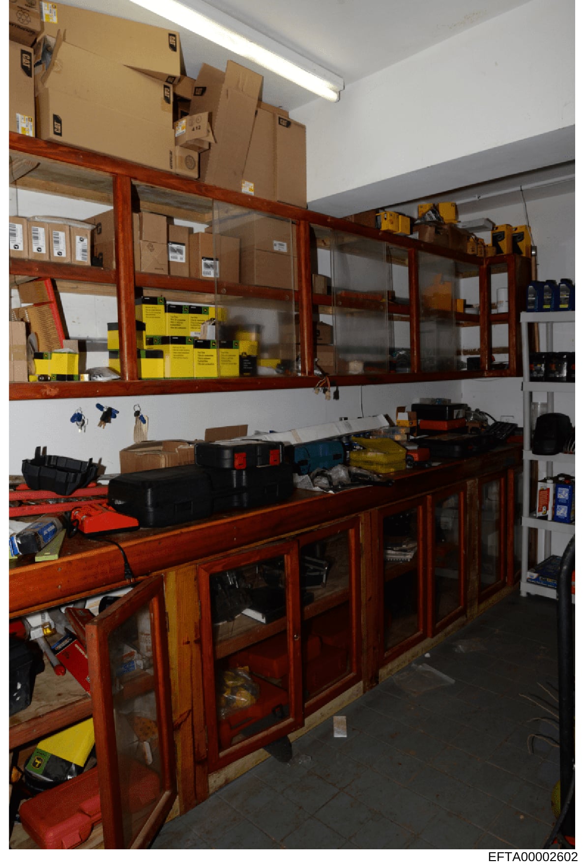 This is a photograph of an organized storage or maintenance room containing various tools, equipment, and supplies stored in wooden cabinets with glass doors and on open shelving. The room features bright yellow warning labels or signage on multiple 