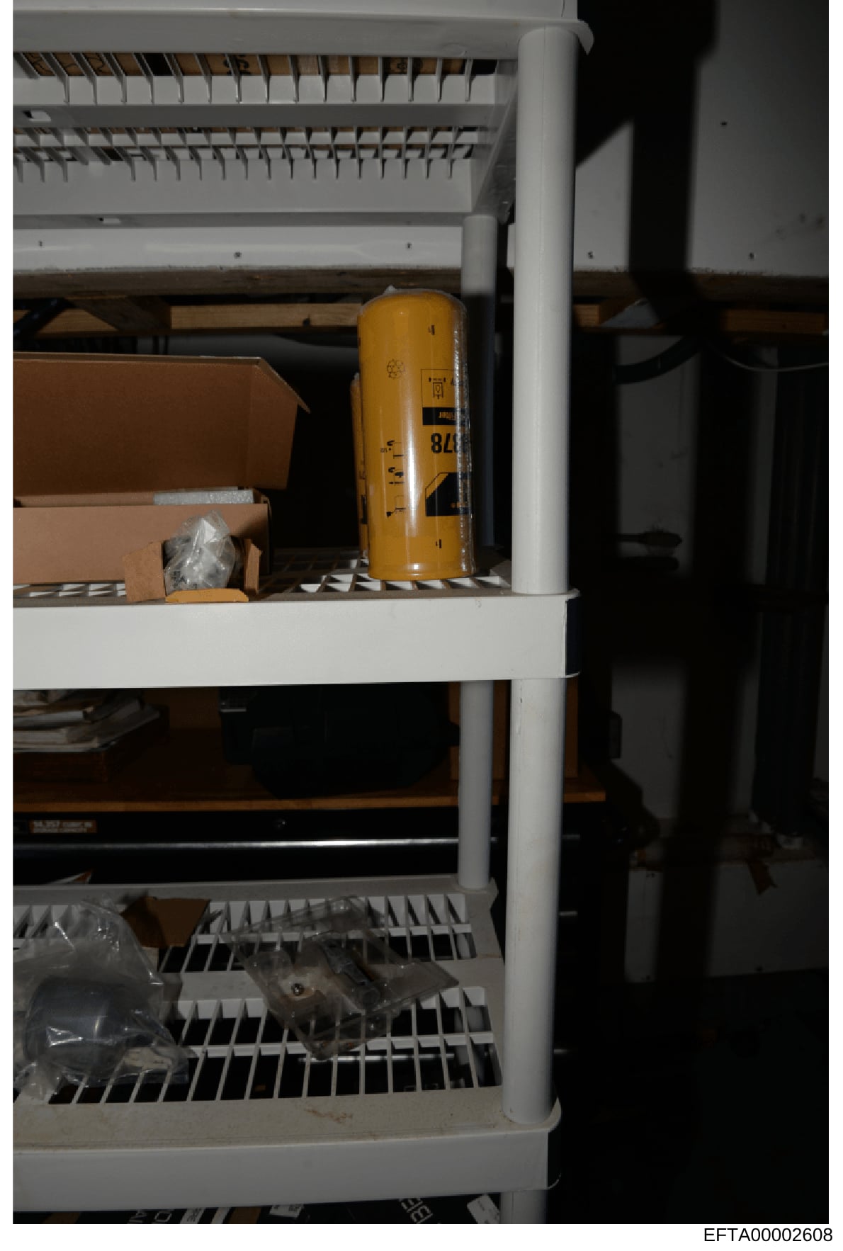 This is an evidence photograph showing a white metal shelving unit containing various stored items including cardboard boxes, a yellow cylindrical container, plastic-wrapped materials, and electrical equipment. The photo appears to be documentation o
