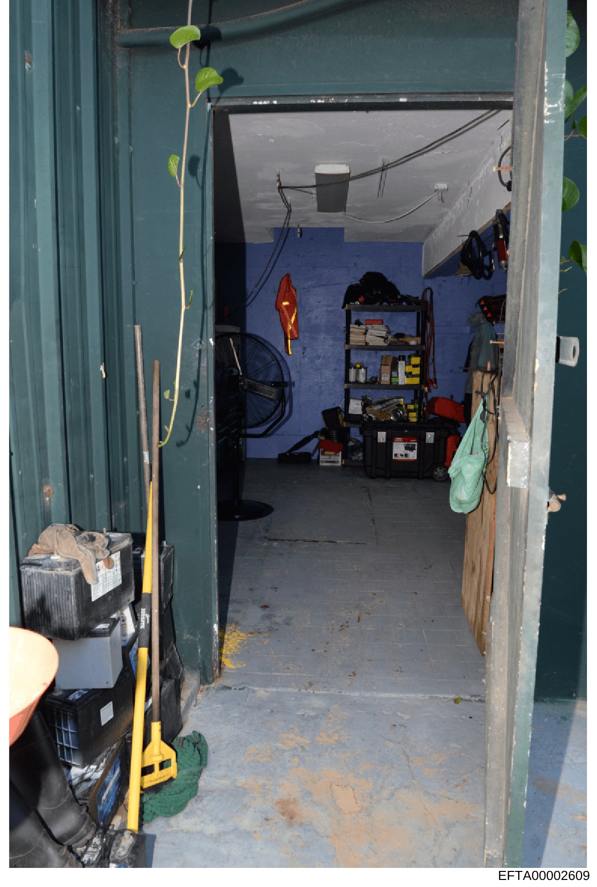 This is a photograph of a storage space or garage interior, showing various tools, equipment, and personal items organized on shelves and the floor. The space has blue flooring, teal/green walls, and contains items such as a red kayak, shelving unit