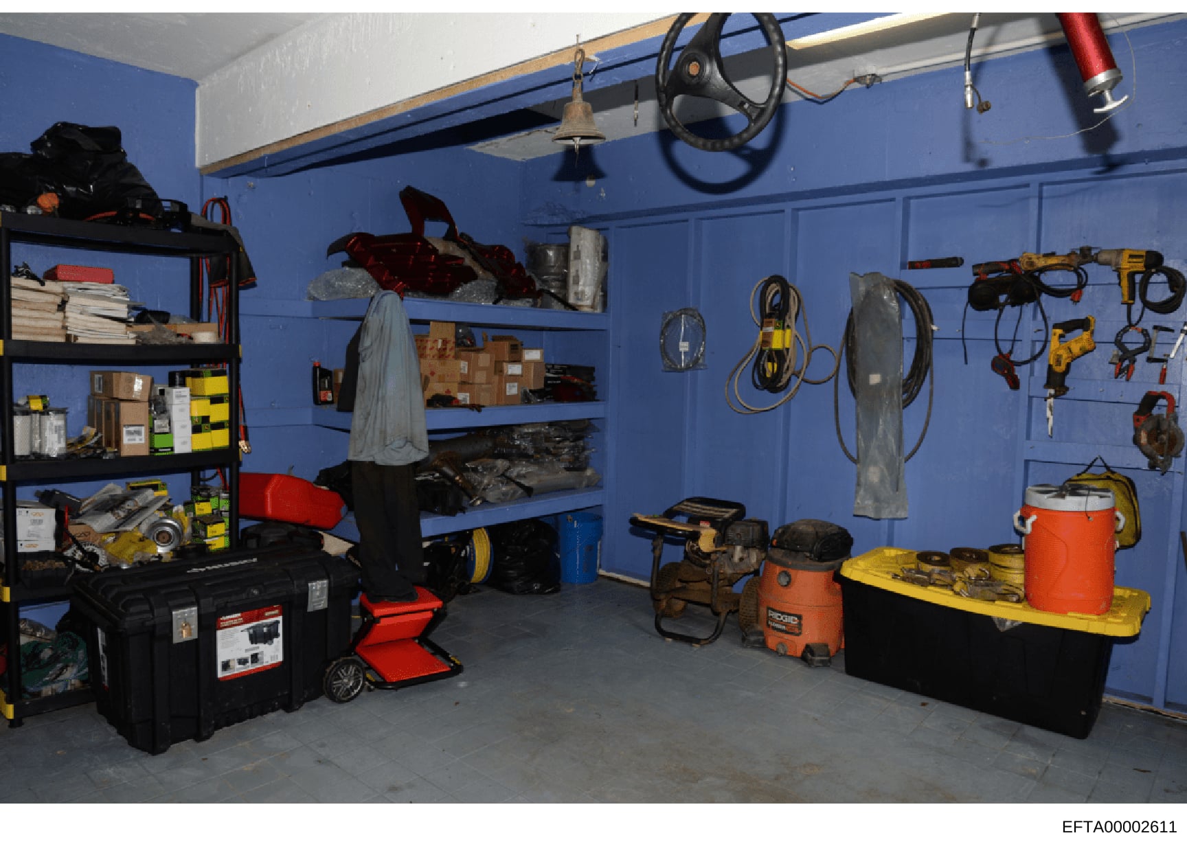 This photograph documents a garage or workshop storage area with bright blue walls, containing various tools, equipment, and supplies. The space includes shelving units, power tools, cords, containers, and miscellaneous items organized on walls and f