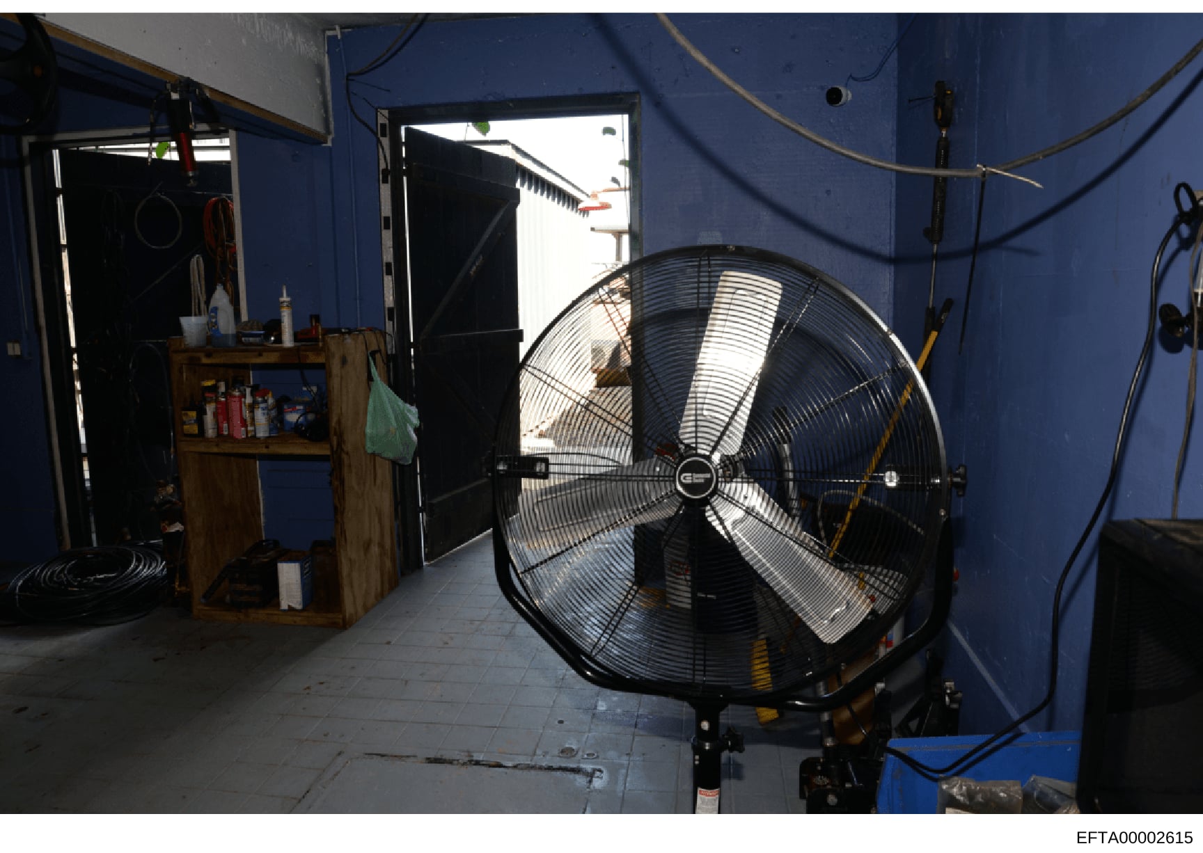 This is an evidence photograph showing the interior of a room with bright blue walls, containing a large industrial-grade fan, wooden storage cabinet with supplies, and various equipment and cords. The image appears to be documentation of a property 