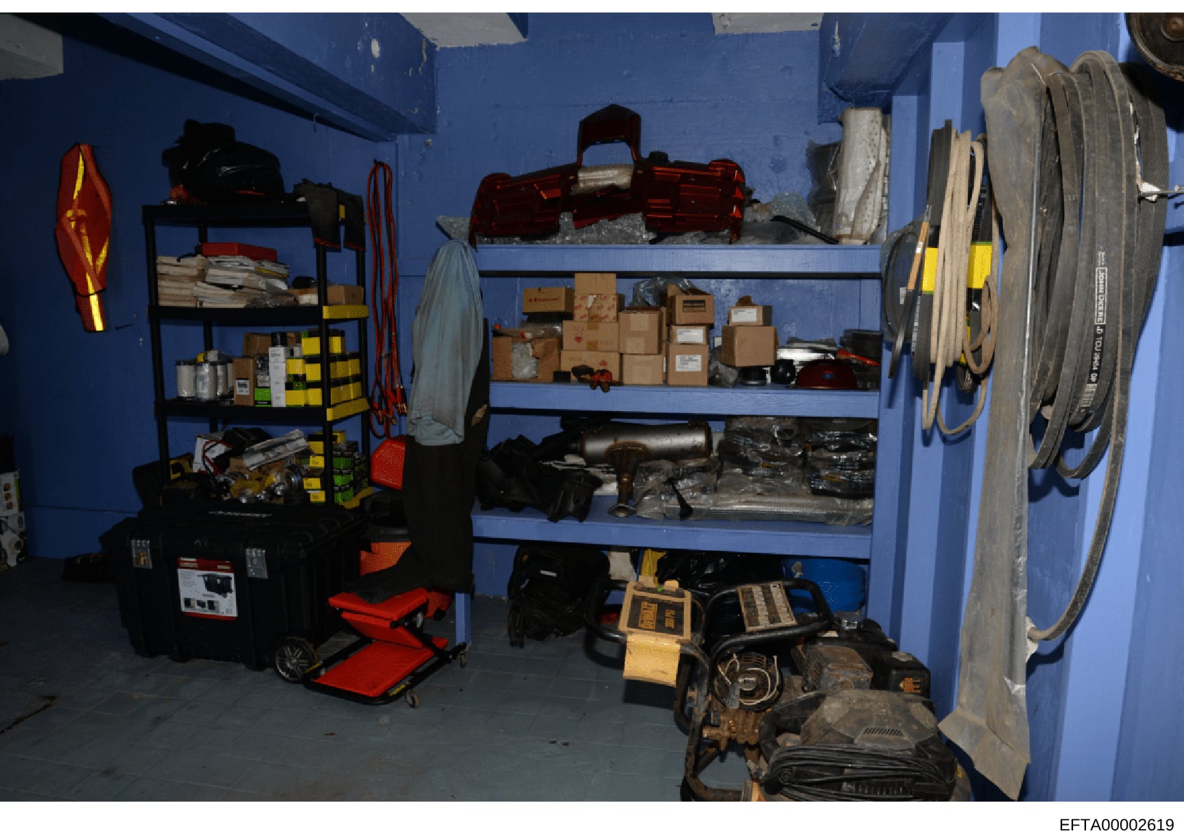 This is a photograph of a storage or utility room with bright blue walls containing various tools, equipment, and supplies. The room includes metal shelving units with cardboard boxes, automotive parts, electrical equipment, coils of wire or rope, an