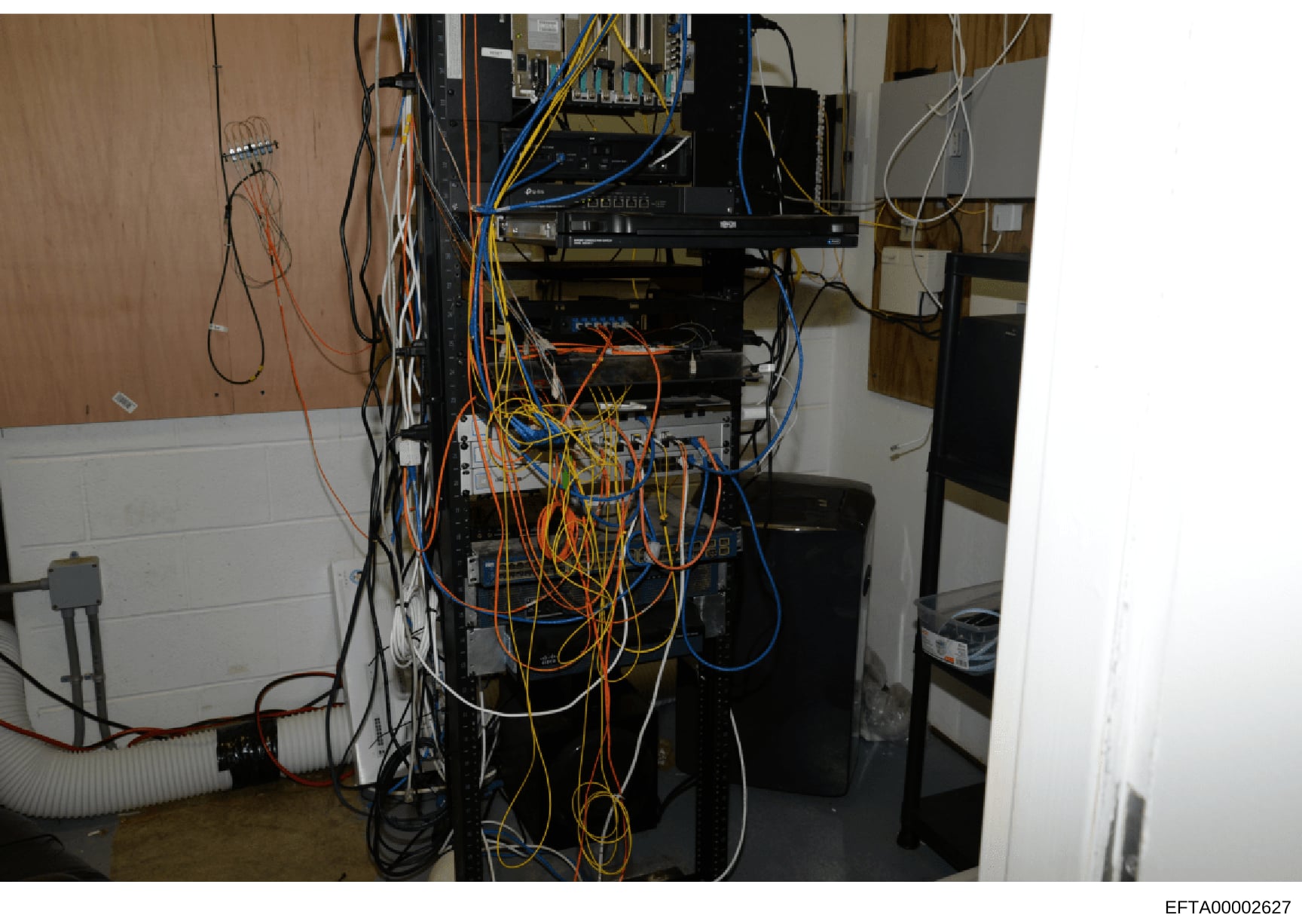 This photograph depicts a wall-mounted electrical and networking infrastructure installation, featuring multiple servers, circuit breakers, and extensive cabling in orange, blue, and yellow. The image appears to be evidence documentation from a prope