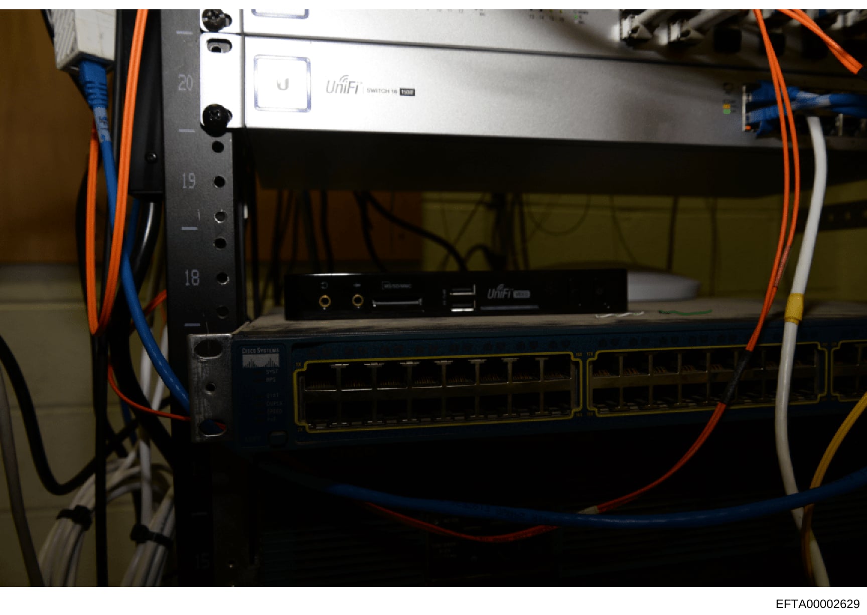 This is a photograph of network infrastructure equipment, showing a rack-mounted setup with UniFi networking equipment, various cables (orange, blue, yellow), and what appears to be server or network switching hardware. The image documents IT infrast