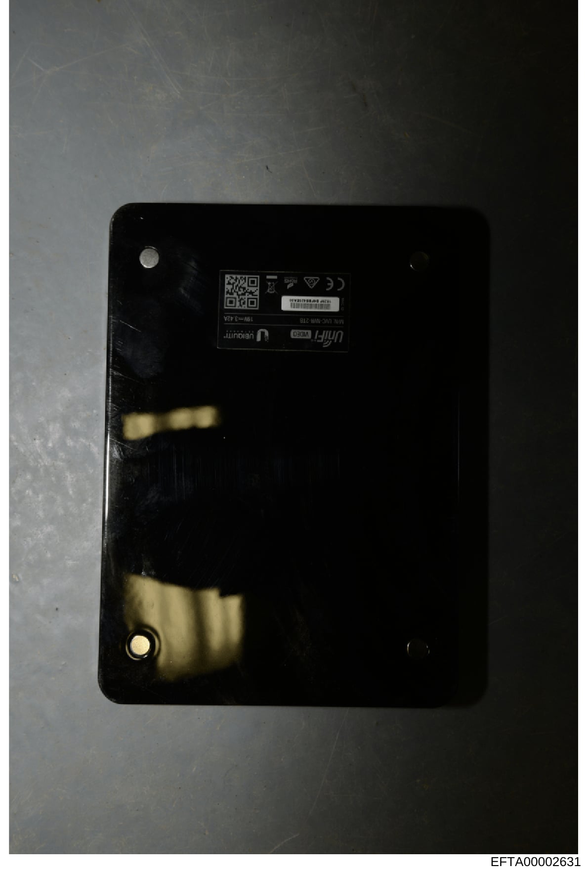 This is a photograph of the back panel of an electronic device, showing regulatory markings, a QR code, and device identification information. The image appears to be evidence documentation, with visible wear and discoloration on the black surface. T