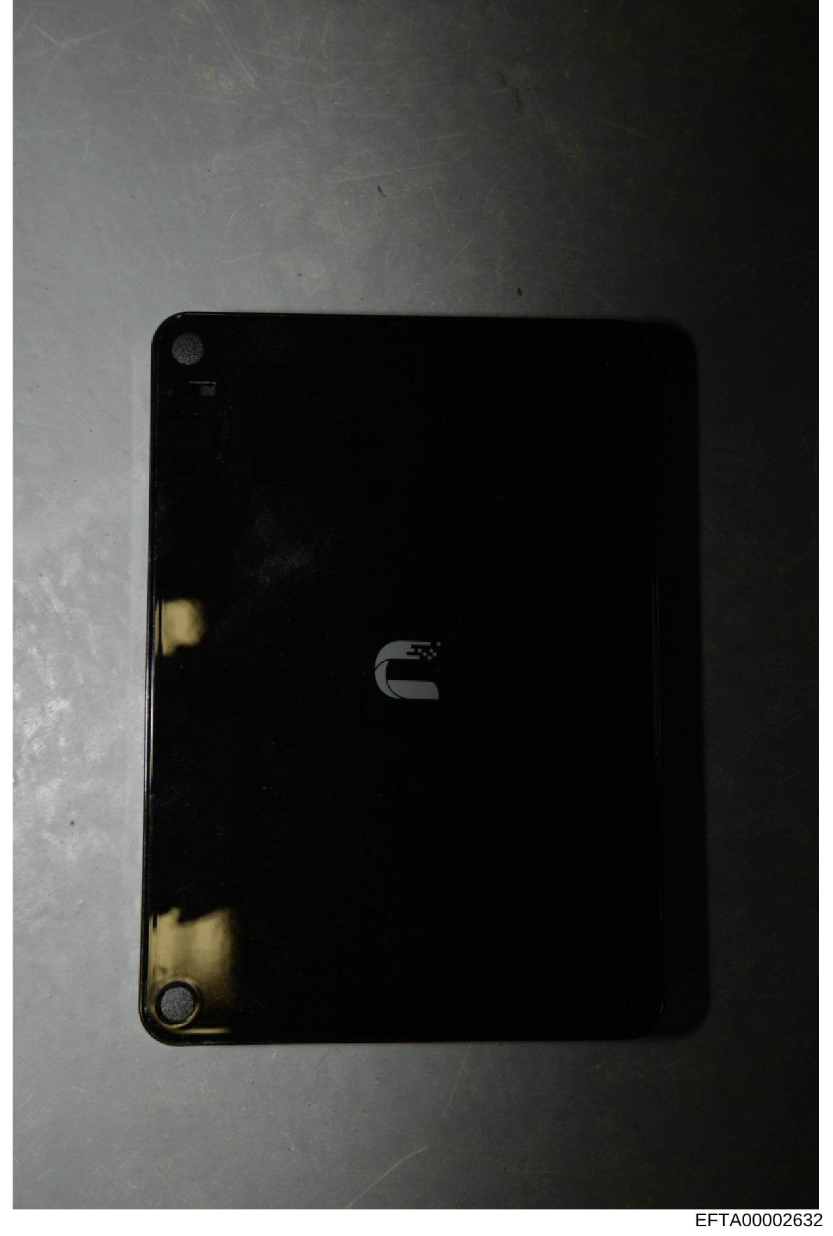 This is a photograph of a black electronic device, likely a computer or laptop, shown from above against a light background. The device displays a logo on its back cover and shows signs of wear and age. This appears to be evidence documentation from