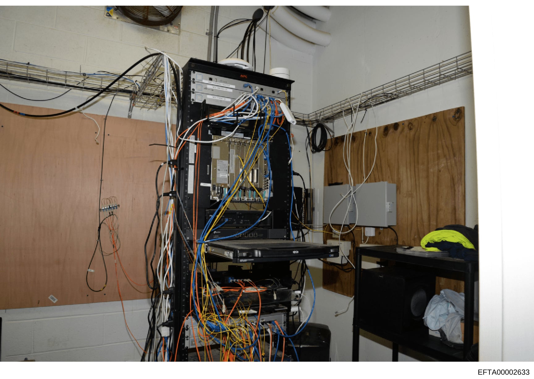 This photograph depicts a secured server room or network equipment facility containing a black server rack with multiple pieces of networking and computing hardware, extensive cable management systems, and mounted control panels. The image shows acti
