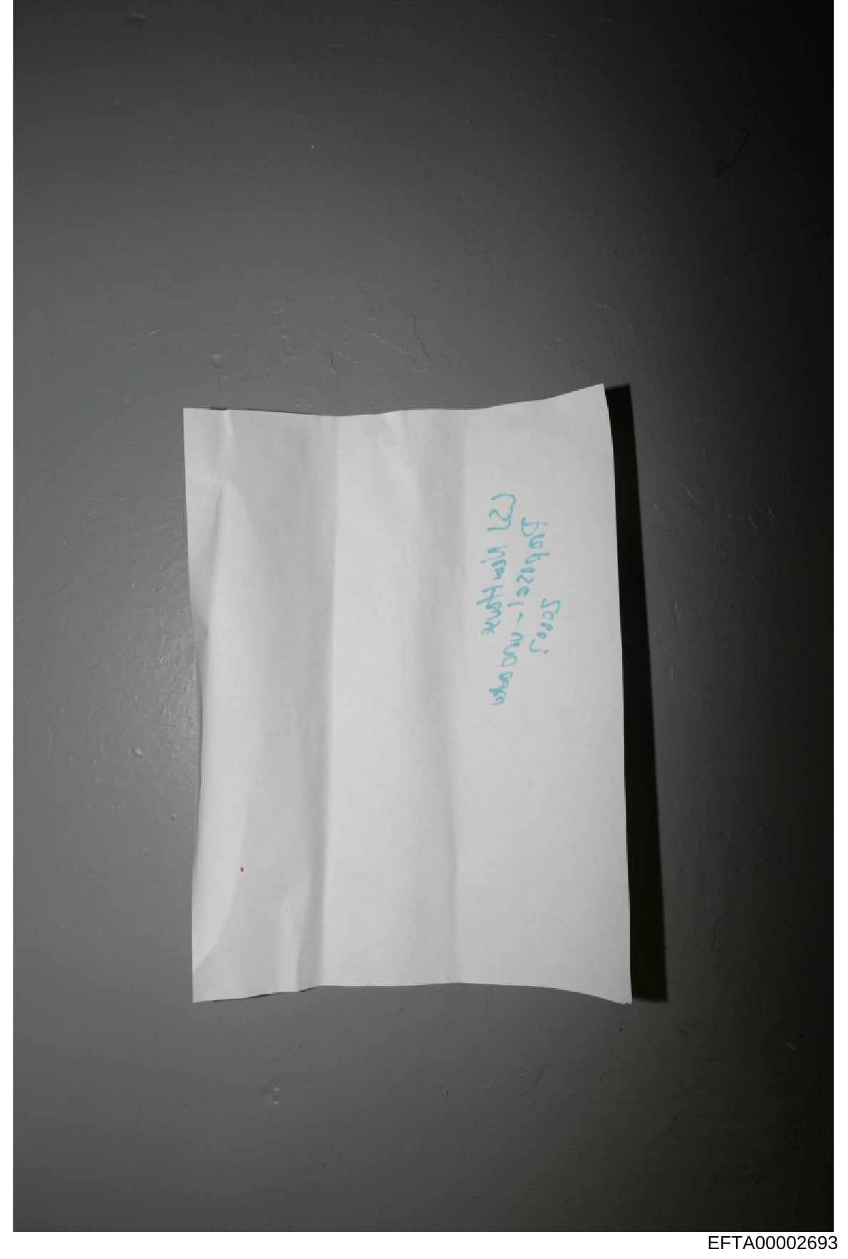 This is a photograph of a white folded piece of paper with handwritten text in blue ink. The handwriting is difficult to read clearly in the image, but appears to contain cursive writing. The document is photographed against a dark gray background. W