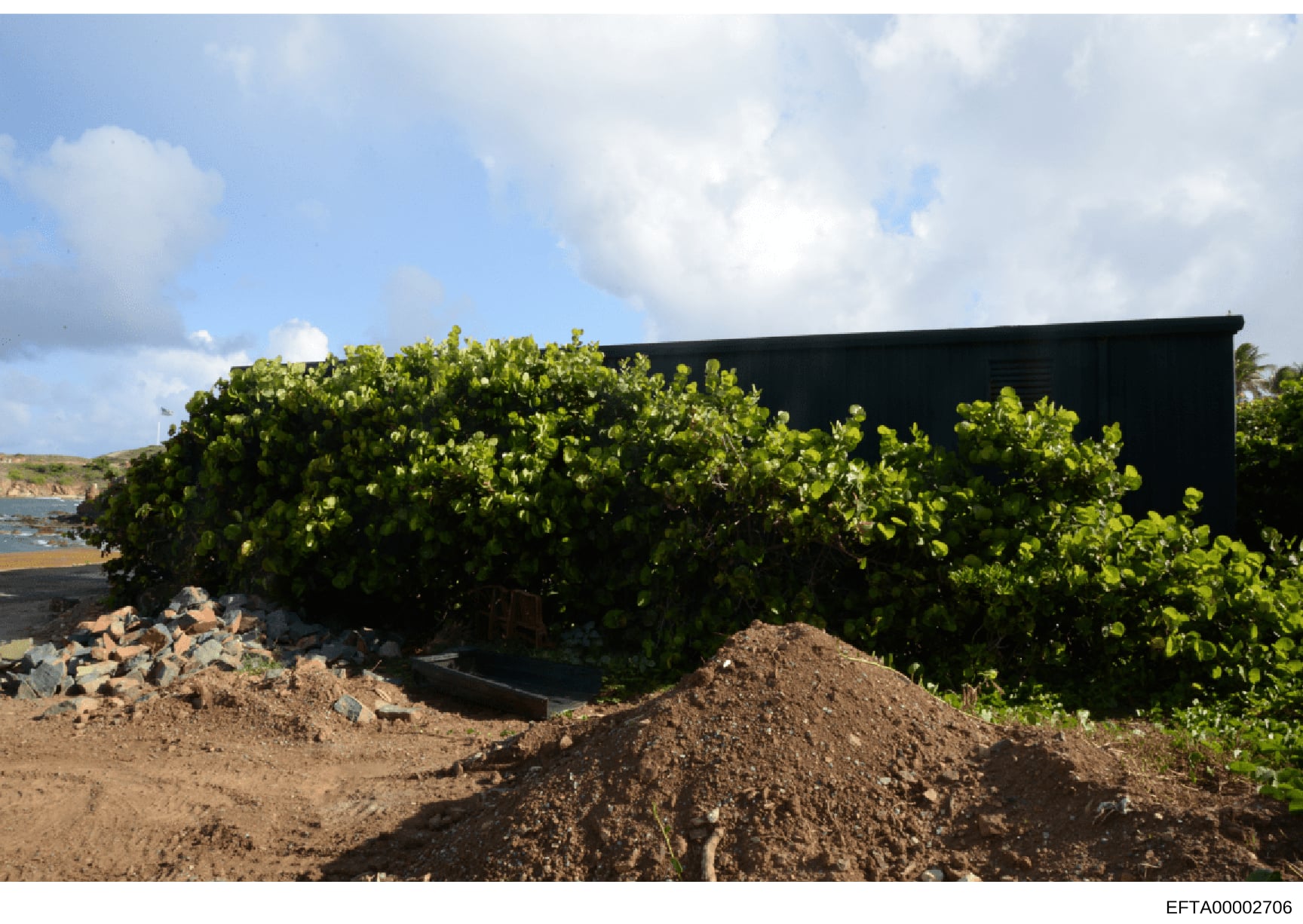 This is a photograph of a black-roofed structure located on a coastal property, partially obscured by dense green vegetation and mangroves. The building sits on sandy/rocky terrain with ocean and cliff views visible in the background. The image appea