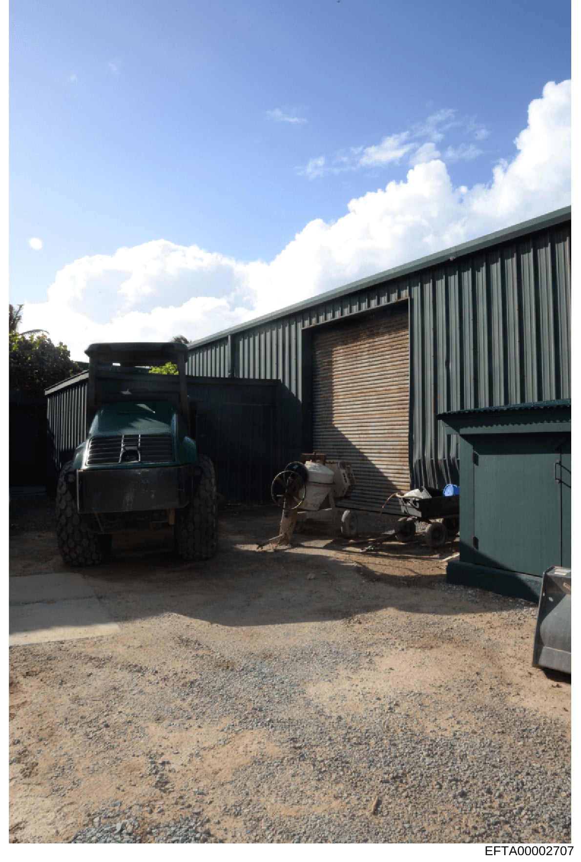 This is a photograph of an industrial or agricultural storage compound featuring a large metal-sided warehouse building with corrugated metal construction. A dark truck is parked in front of the facility, with various equipment and storage containers