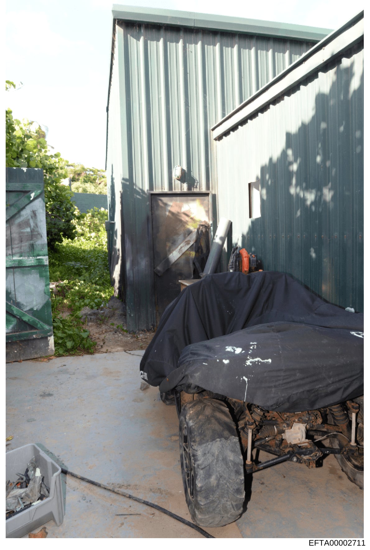 This photograph shows an outdoor storage or work area adjacent to a metal-sided building. The image captures a covered piece of equipment or machinery under black tarps, along with various stored items and debris. The presence of overgrown vegetation