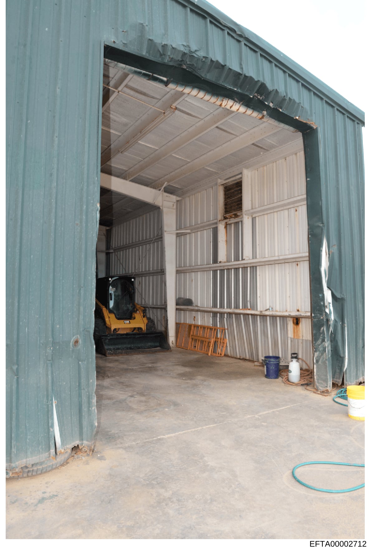 This is an evidence photograph showing the interior of a large industrial metal building with corrugated siding. The space appears to be a storage facility or garage, containing a yellow and black piece of machinery (possibly a skid steer loader or s