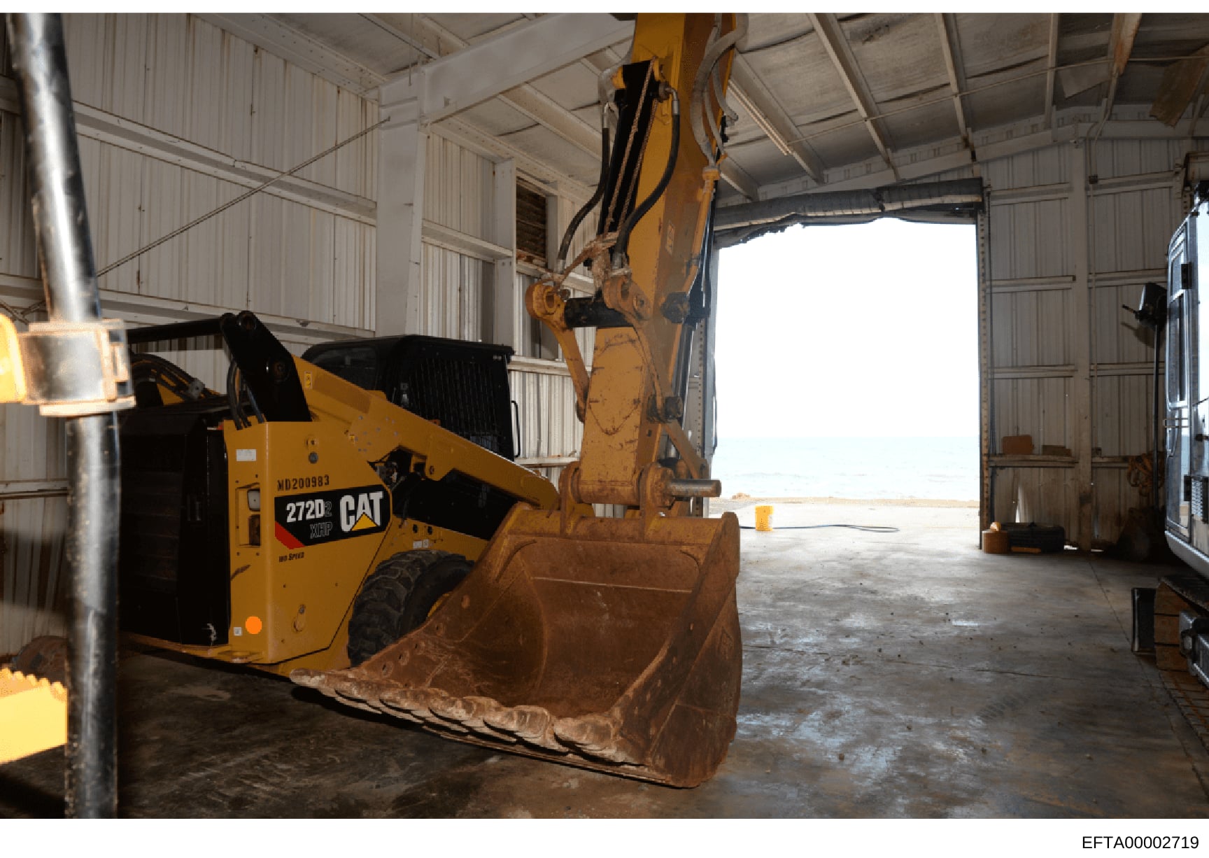 This photograph shows a large yellow Caterpillar 272D skid steer loader (Model WD200983) parked inside a metal corrugated warehouse or storage facility. The equipment appears to be in an industrial or construction yard setting. The relevance to the E
