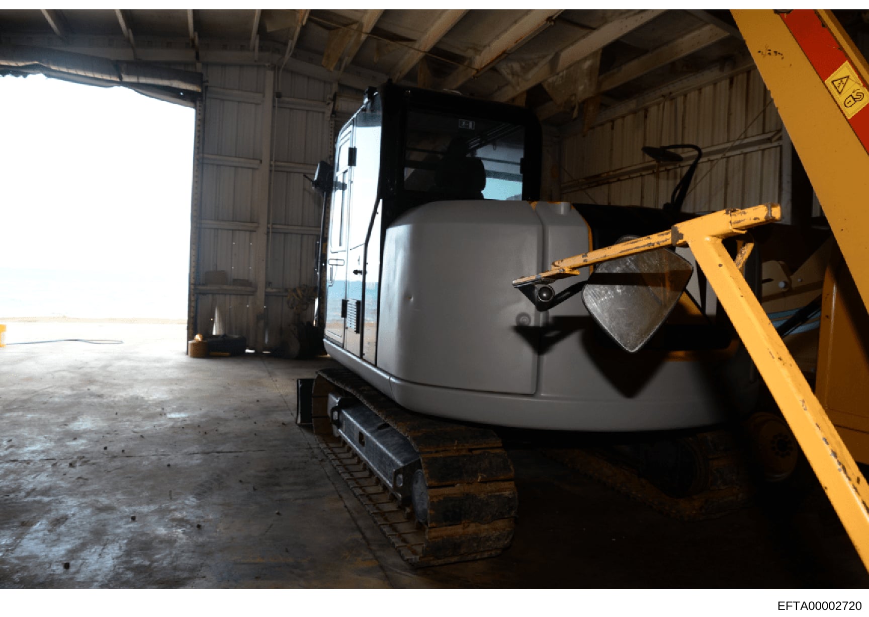 This photograph shows a large piece of heavy construction equipment (excavator) and a white trailer stored inside an industrial warehouse or hangar facility. The image appears to be documentary evidence of seized or inventoried assets, likely taken d