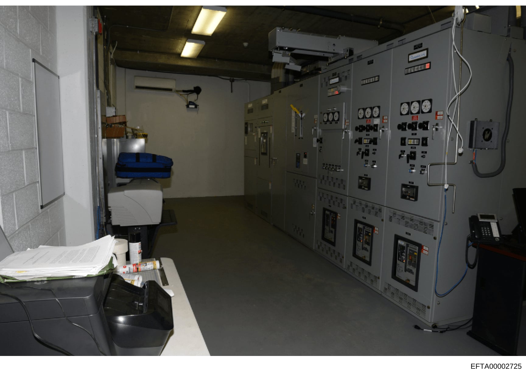 This photograph depicts an industrial or commercial electrical control room featuring large gray electrical panels with monitoring and control systems. A work desk with documents and supplies is visible in the foreground, suggesting an operational mo
