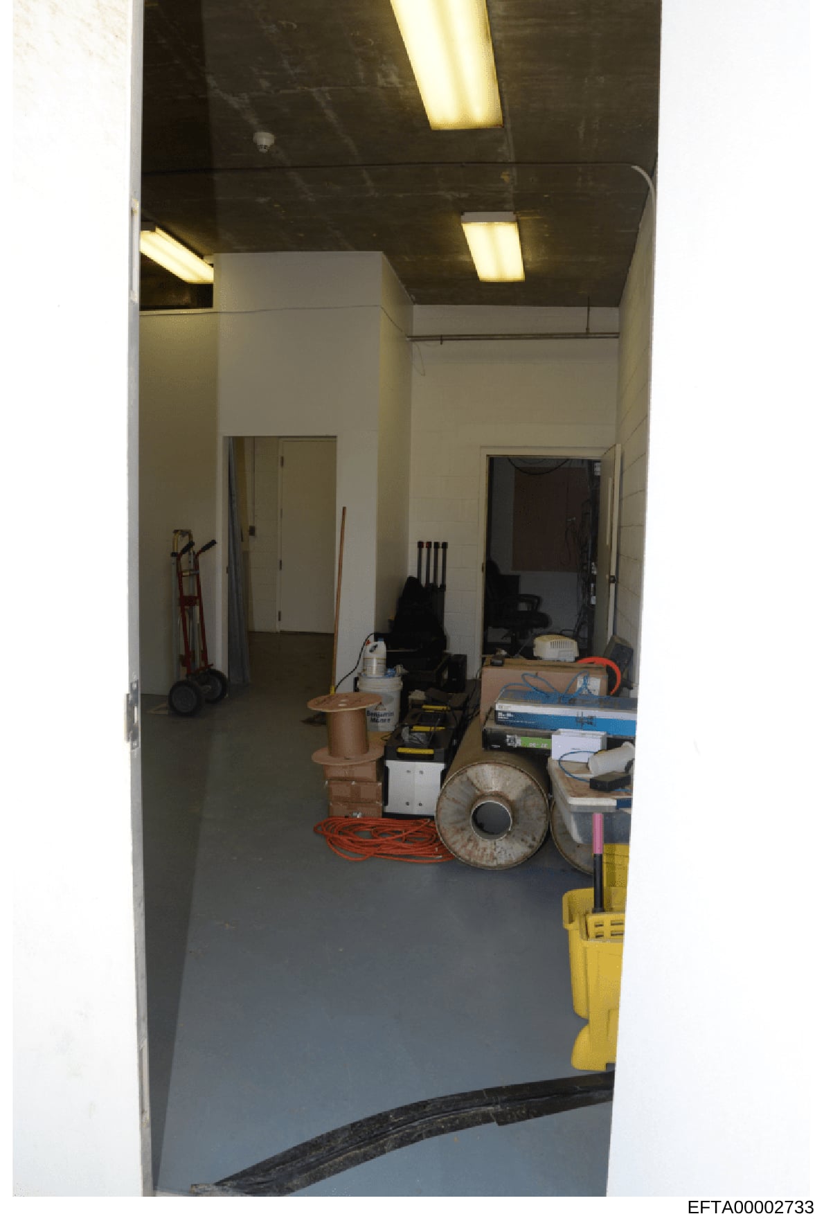 This is a photograph of an interior utility or storage room containing various maintenance and construction equipment. The space appears to be part of a larger property under investigation, with fluorescent ceiling lighting, white walls, and concrete