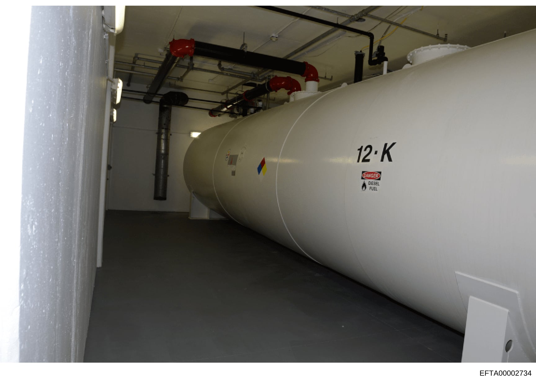 This photograph documents a large-capacity diesel fuel storage tank (labeled '12·K') installed in an underground or basement mechanical room. The tank is equipped with safety piping, red fire suppression equipment mounted on the ceiling, and proper h