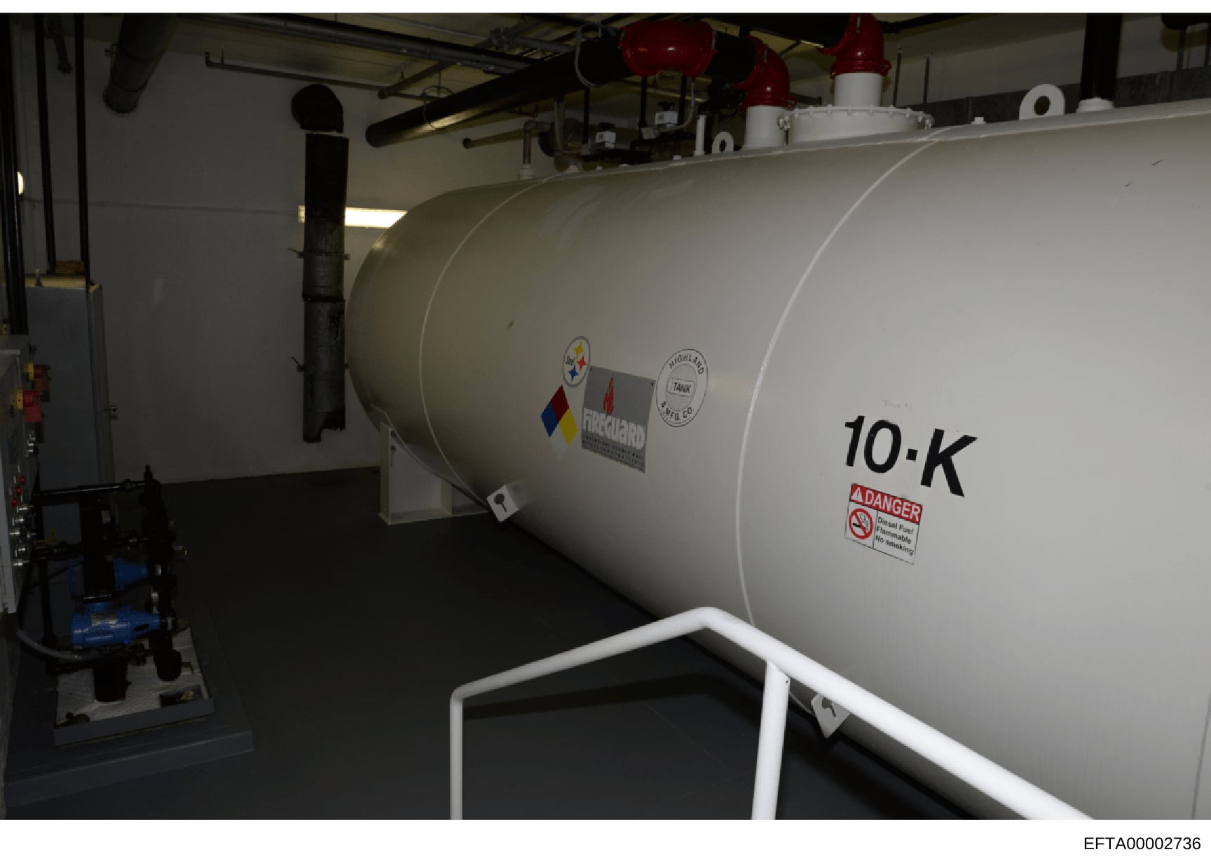 Photograph of a large white cylindrical industrial storage tank labeled '10-K' with a danger warning label, located inside a warehouse or facility. The tank appears to be a propane or fuel storage container with regulatory markings and safety signage
