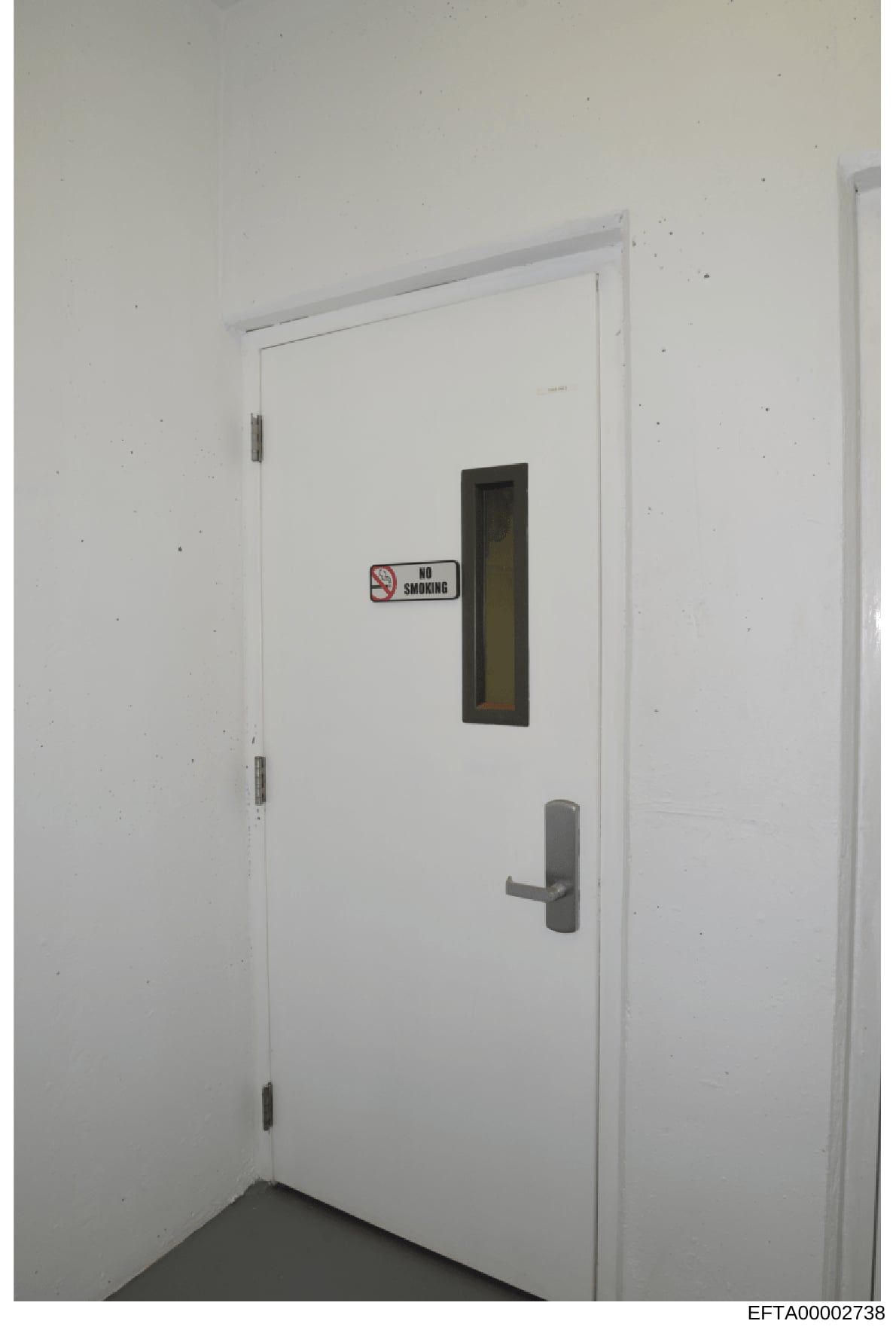 This is a photograph of an interior doorway, likely from one of Epstein's properties. The door is white with a metal handle, hinges, and a small window with dark frame. A standard 'No Smoking' sign is affixed to the door. The image appears to be evid