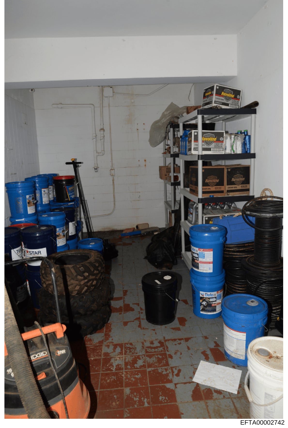 This is a photograph of an interior storage space containing multiple drums of automotive fluids (including Havoline brand products), tires, tools, and maintenance equipment. The room appears to be a garage or maintenance facility storage area, with 
