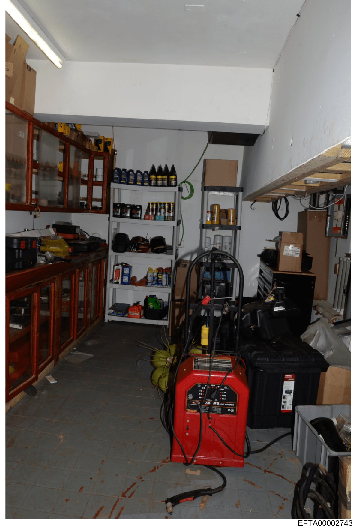 This photograph shows the interior of a storage or utility room, possibly at one of Epstein's properties. The space contains various equipment including a red industrial machine (possibly a dehumidifier or power tool), wooden cabinets with glass door