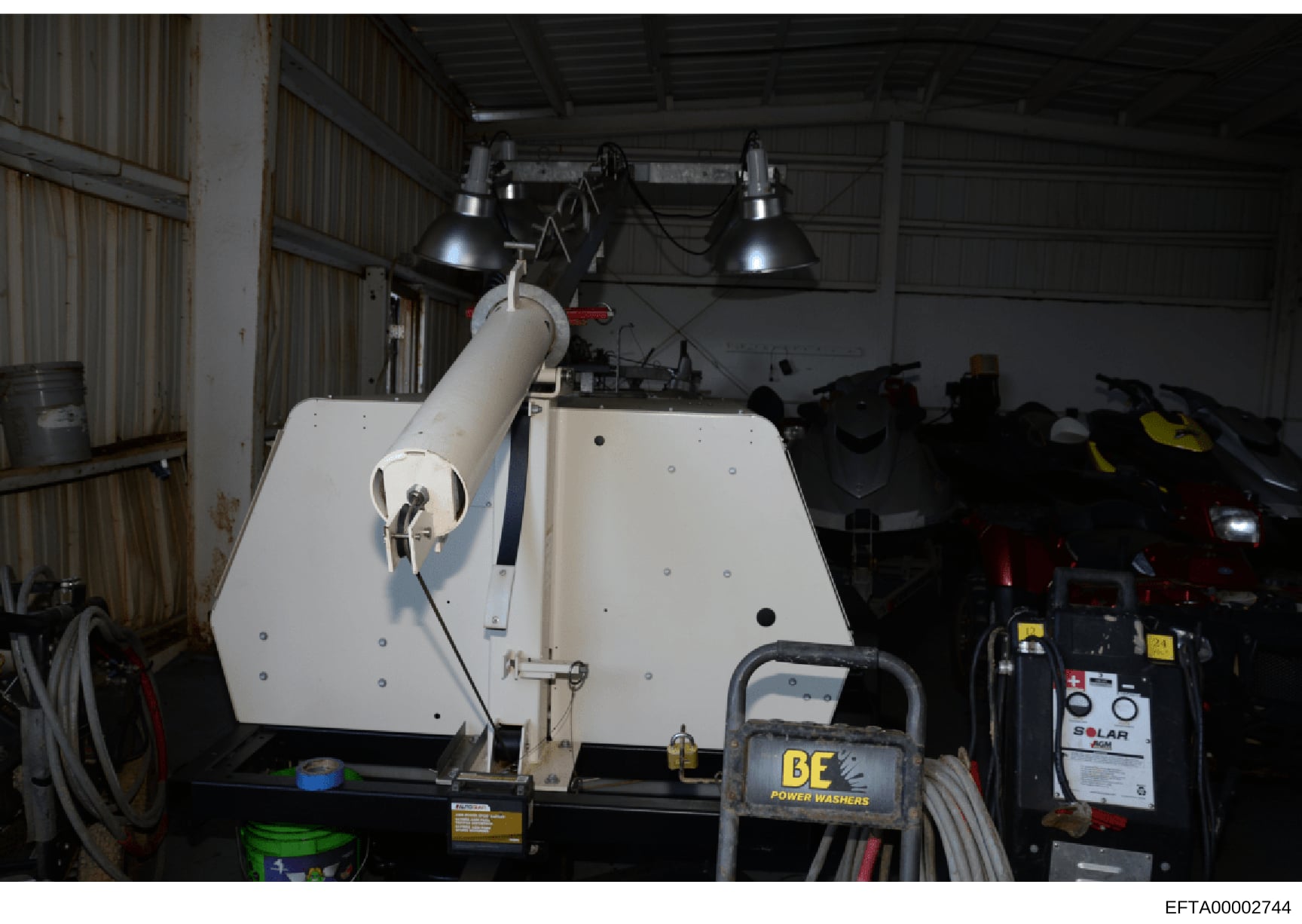 This photograph depicts the interior of an industrial warehouse or storage facility containing various equipment and machinery. The image shows a white industrial apparatus or tank suspended under overhead lighting, along with pressure washing equipm