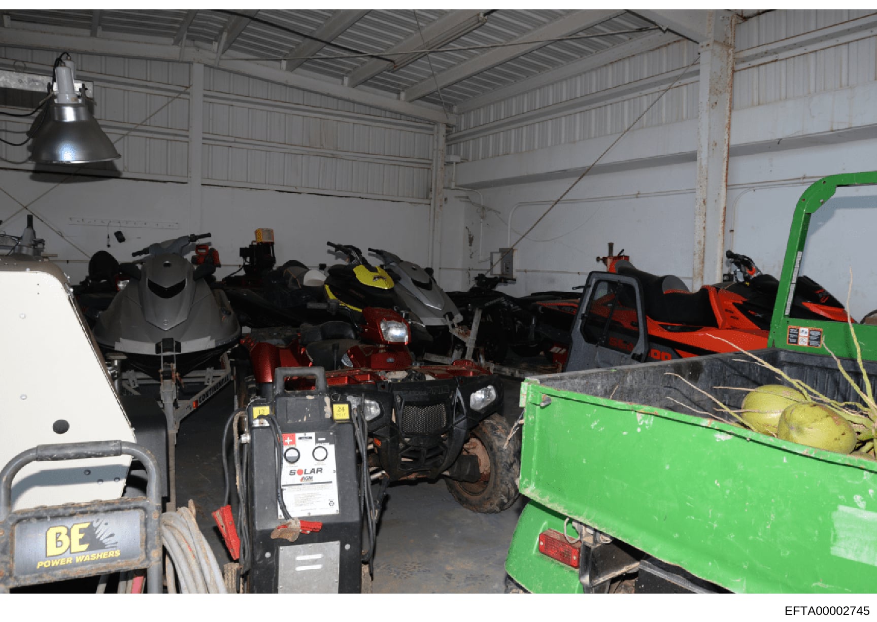This photograph shows the interior of a large metal warehouse or storage facility containing multiple personal watercraft (jet skis), ATVs, power washing equipment, and other recreational vehicles and tools. The image appears to be documentation of a