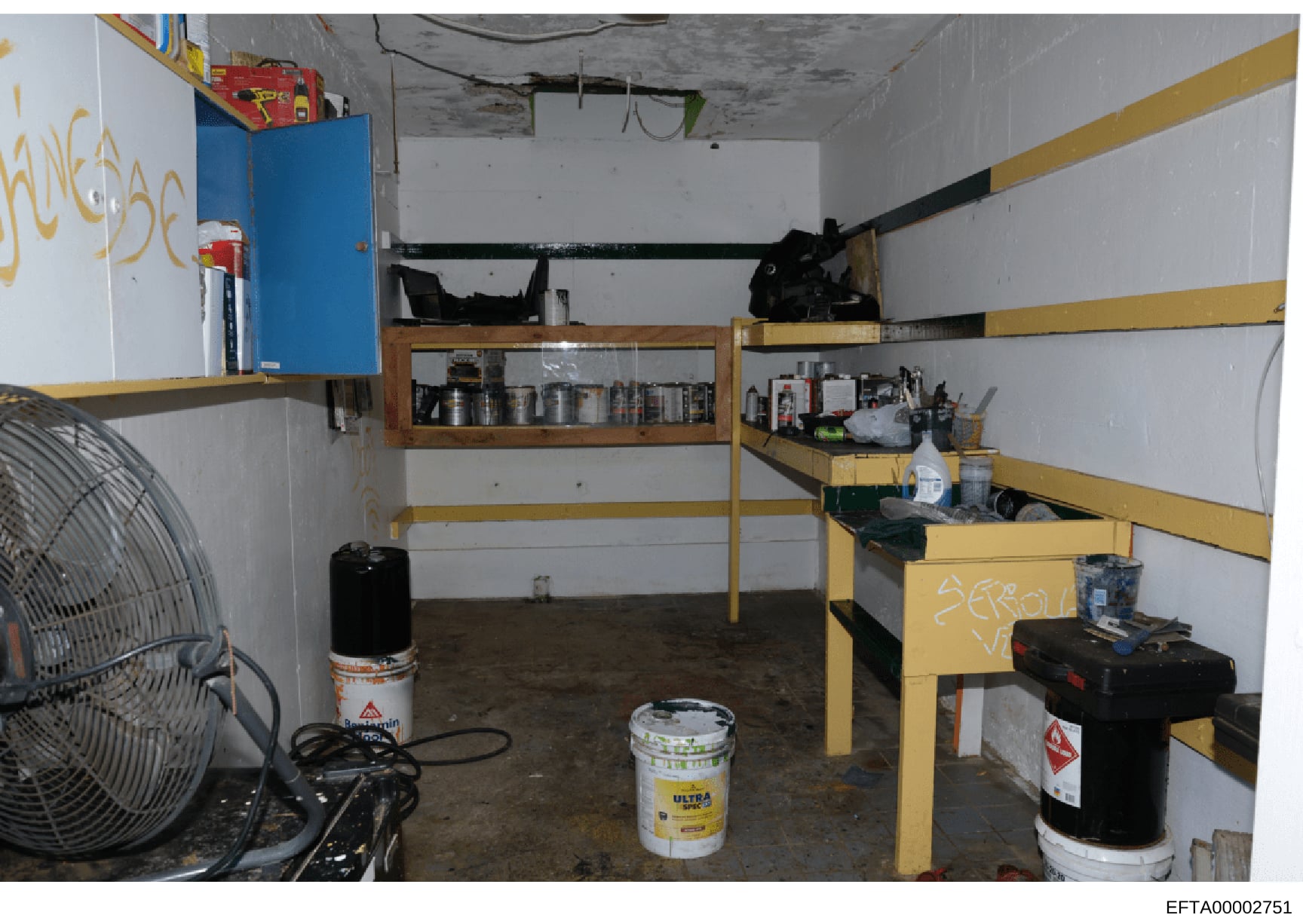 This photograph documents an interior storage or basement room containing yellow metal shelving units, various maintenance and chemical supplies, tools, and equipment. The space appears to be part of a property inventory or evidence documentation, sh