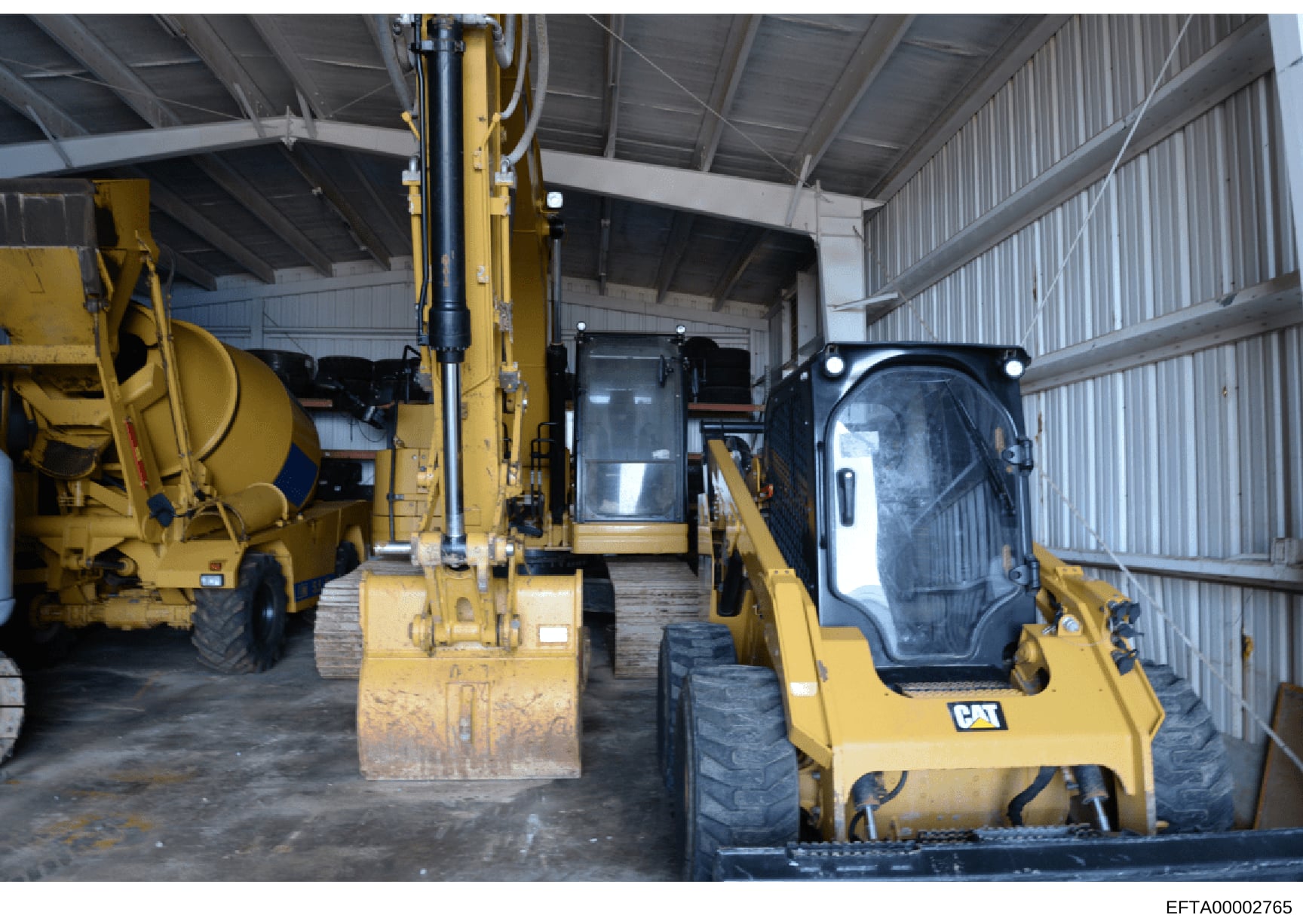 This photograph shows the interior of a large industrial storage facility or warehouse containing heavy construction equipment. The image depicts multiple yellow Caterpillar (CAT) branded pieces of equipment including excavators, a concrete mixer tru
