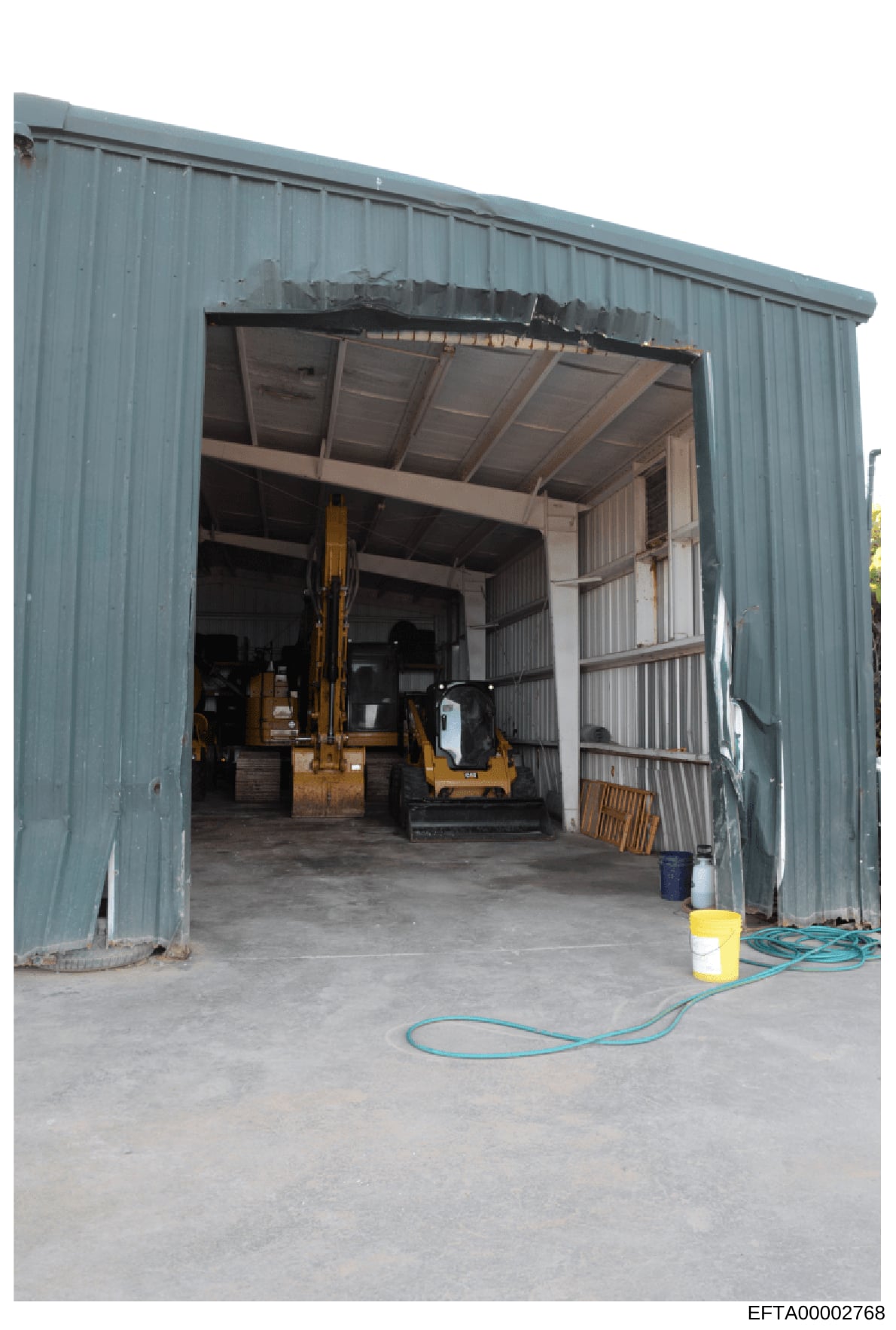 This photograph shows the interior of a large metal-frame warehouse or industrial storage building with corrugated metal siding. The space contains heavy machinery including what appears to be construction or industrial equipment (yellow excavator or