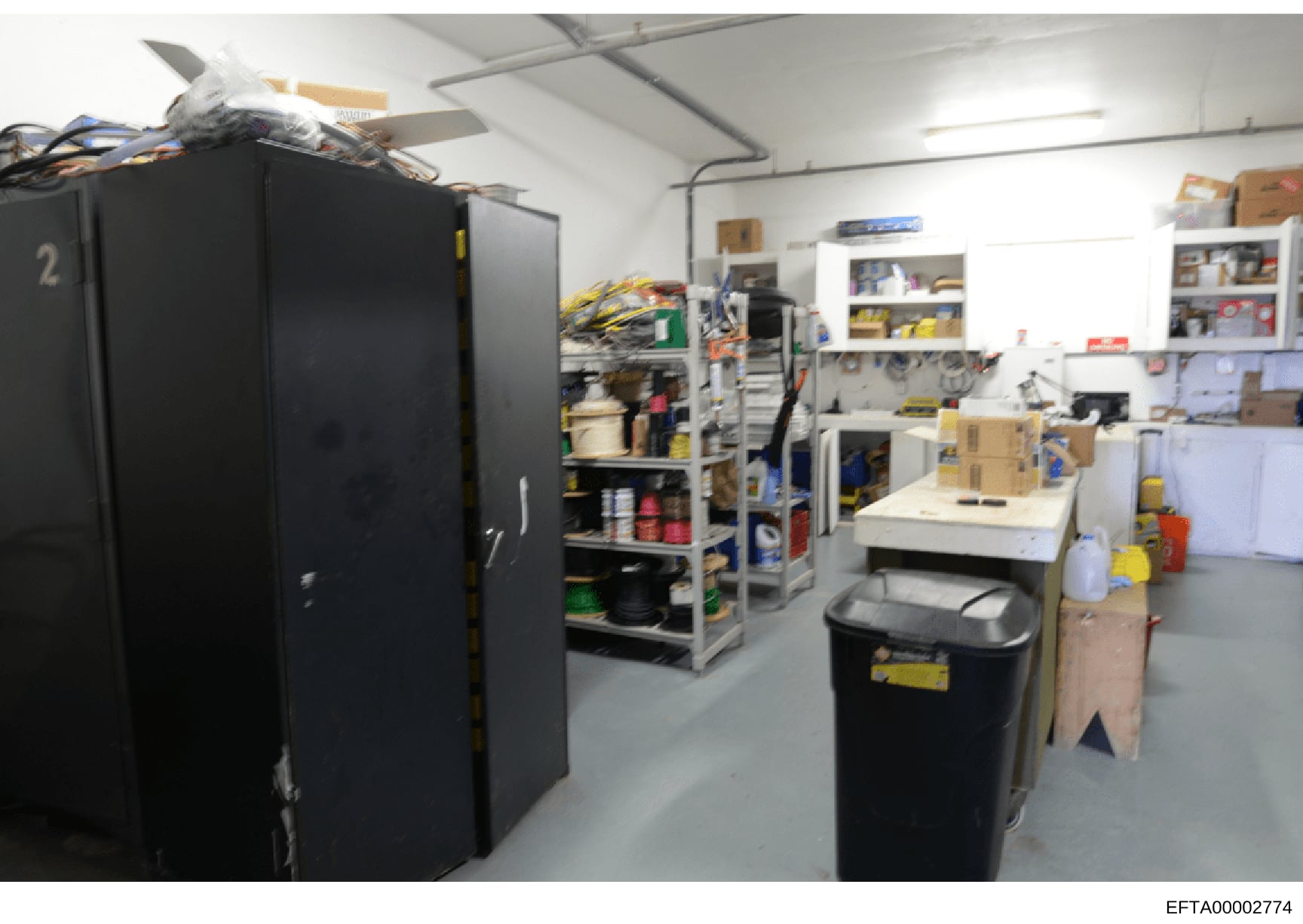 This is a photograph of an industrial or commercial storage facility showing a warehouse-style room with large black cabinet units, metal shelving, work tables, and various supplies and equipment. The room appears to be used for maintenance, storage,
