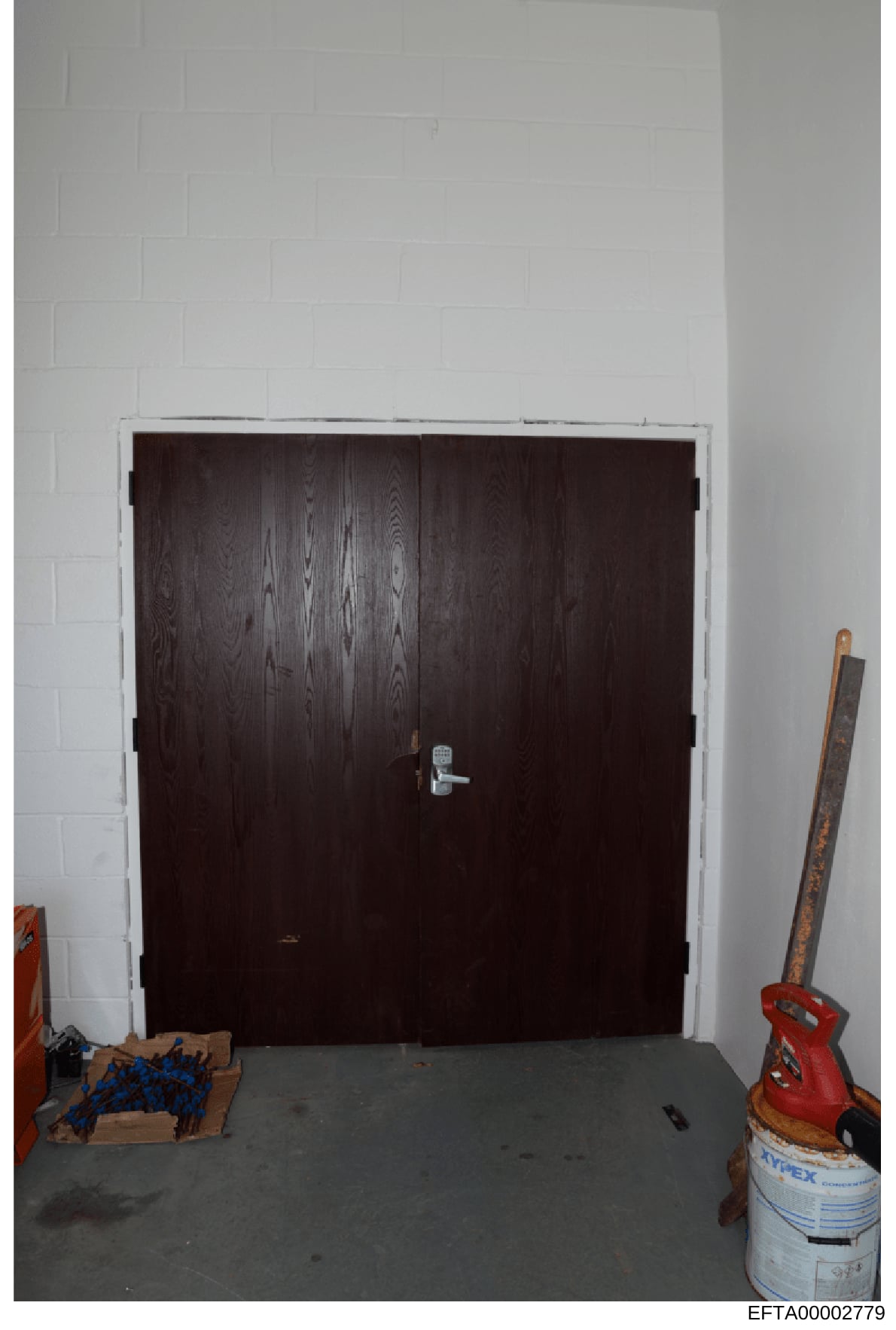This is a photograph of an interior space showing a large dark wooden double-door entry with a metal handle, set in a white cinder block or painted brick wall. The room appears to be under construction or renovation, with various tools and materials