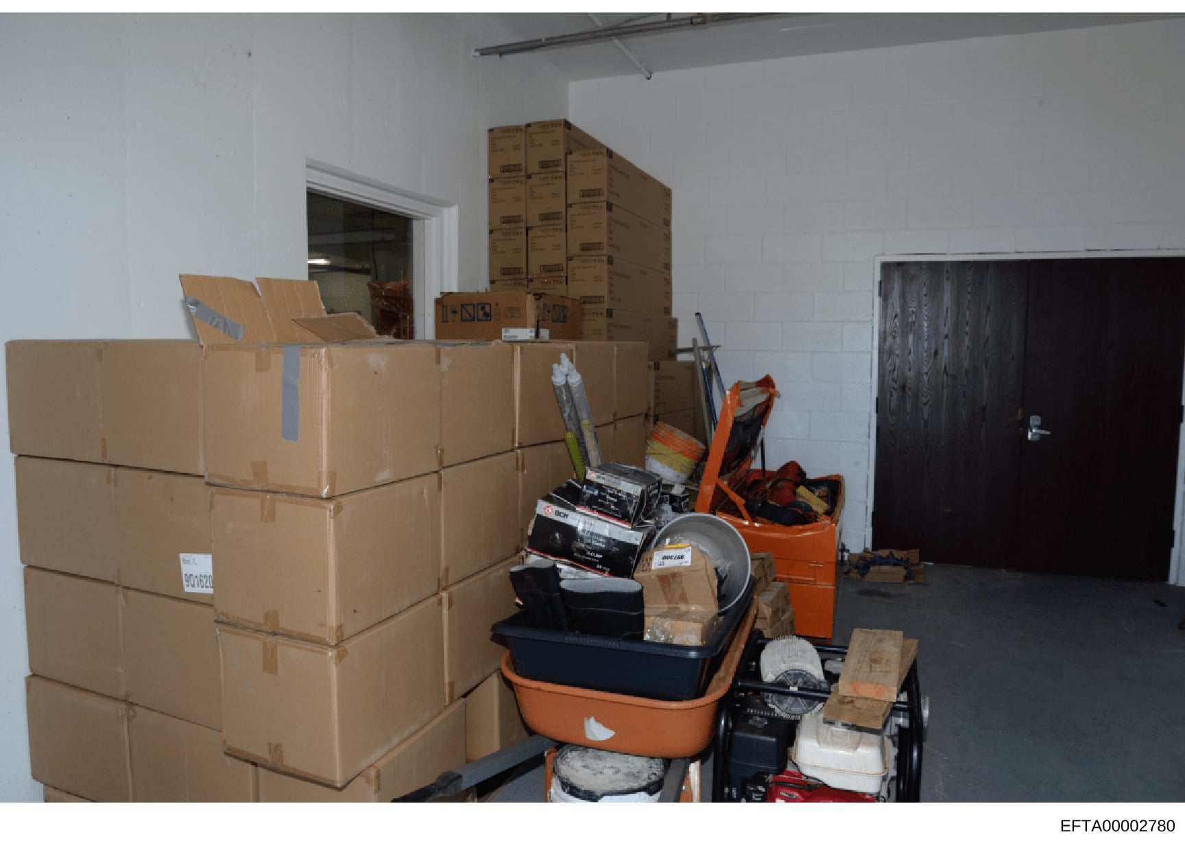 This is an evidence photograph showing a warehouse or storage facility containing numerous sealed cardboard boxes and various household items, tools, and equipment stacked on shelves and the floor. The image appears to be from a property seizure or e