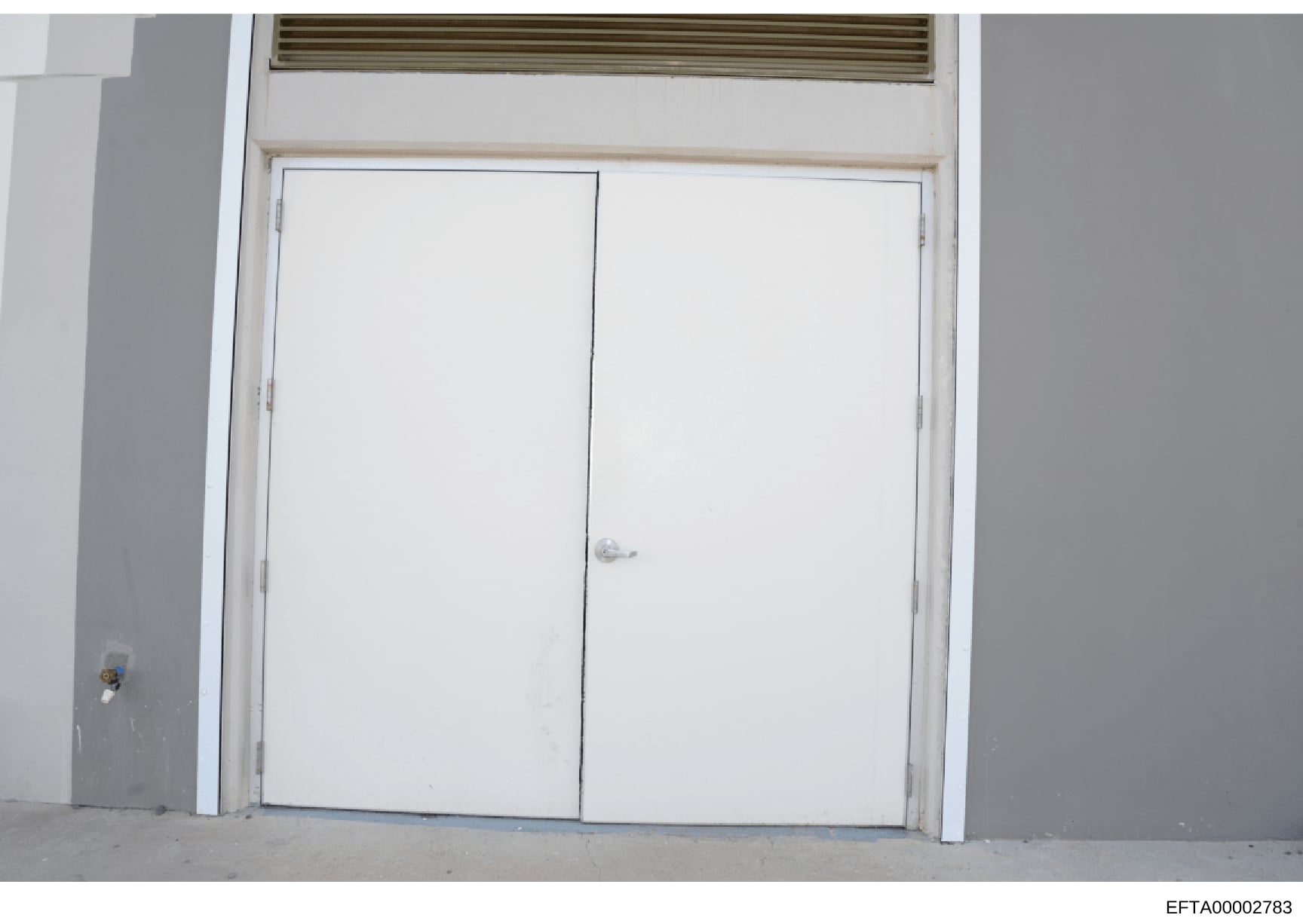 This is a photograph of an interior room showing a set of white double doors with a transom window above. The doors appear to be closet or storage doors in a residential or commercial space. The image shows plain white walls, a concrete floor, and ba