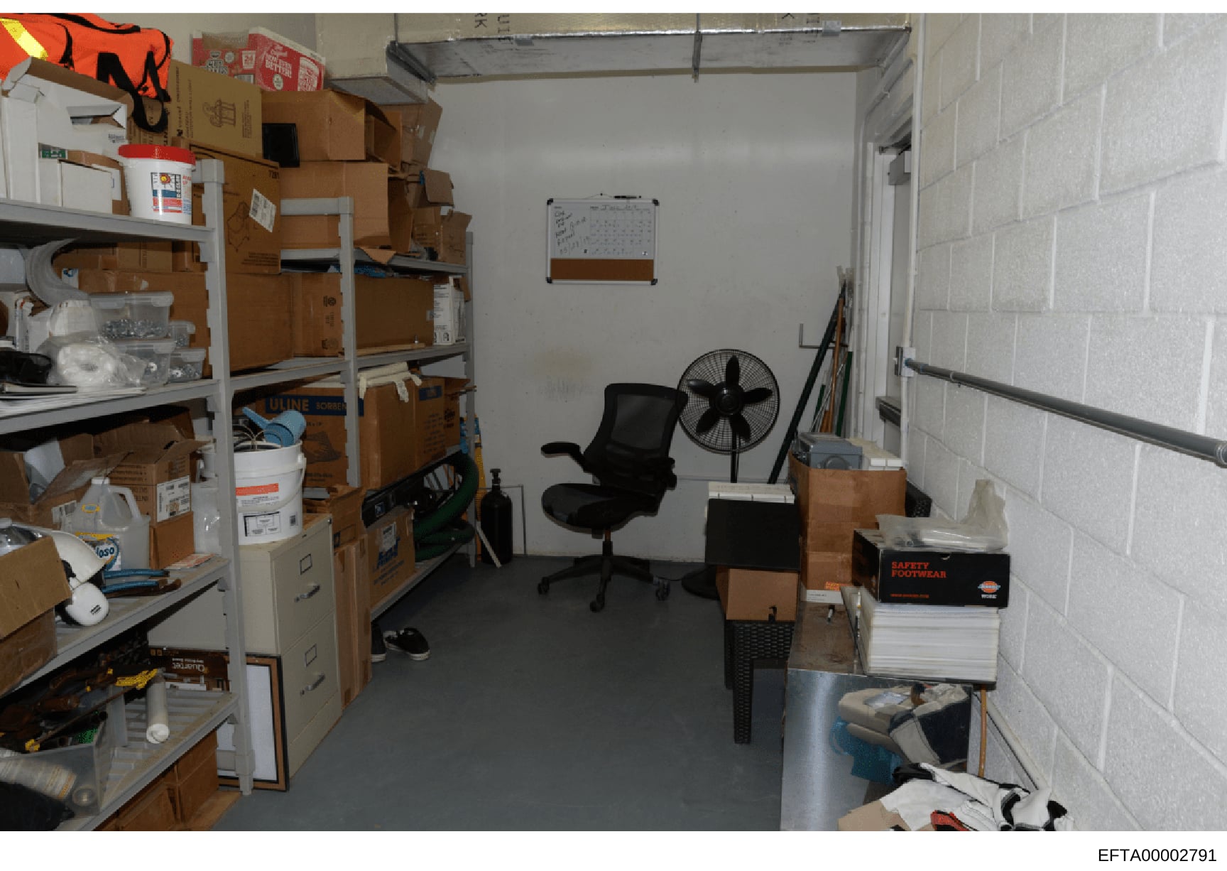 This is an evidence photograph showing the interior of a storage room or basement space containing multiple cardboard boxes, metal shelving units, office furniture, and various supplies. The image appears to be part of a property investigation or ass