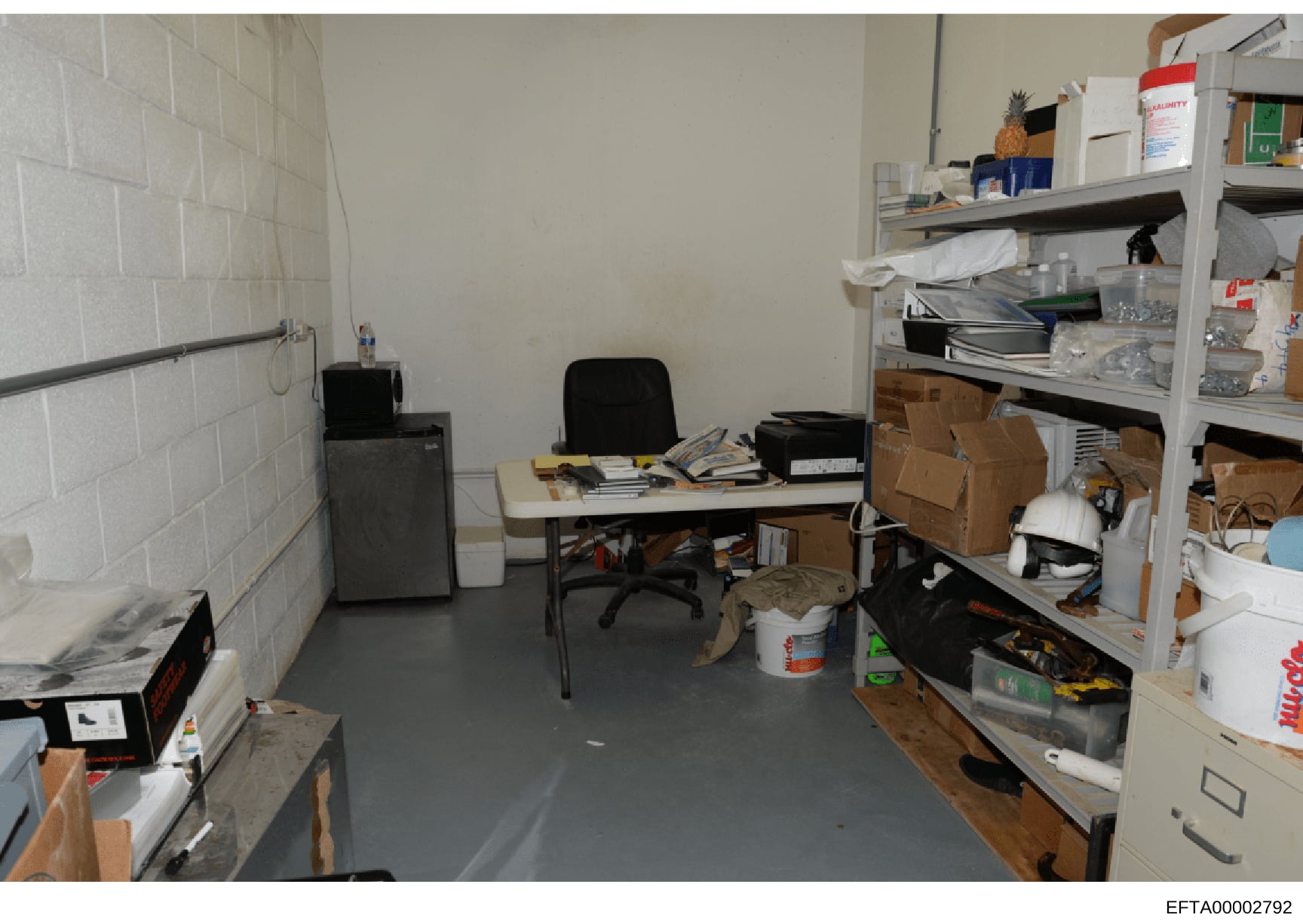 This is an evidence photograph showing an interior room with minimal furnishings and significant clutter. The space contains a desk with office chair, shelving units stocked with boxes and supplies, and various items scattered throughout. The room fe