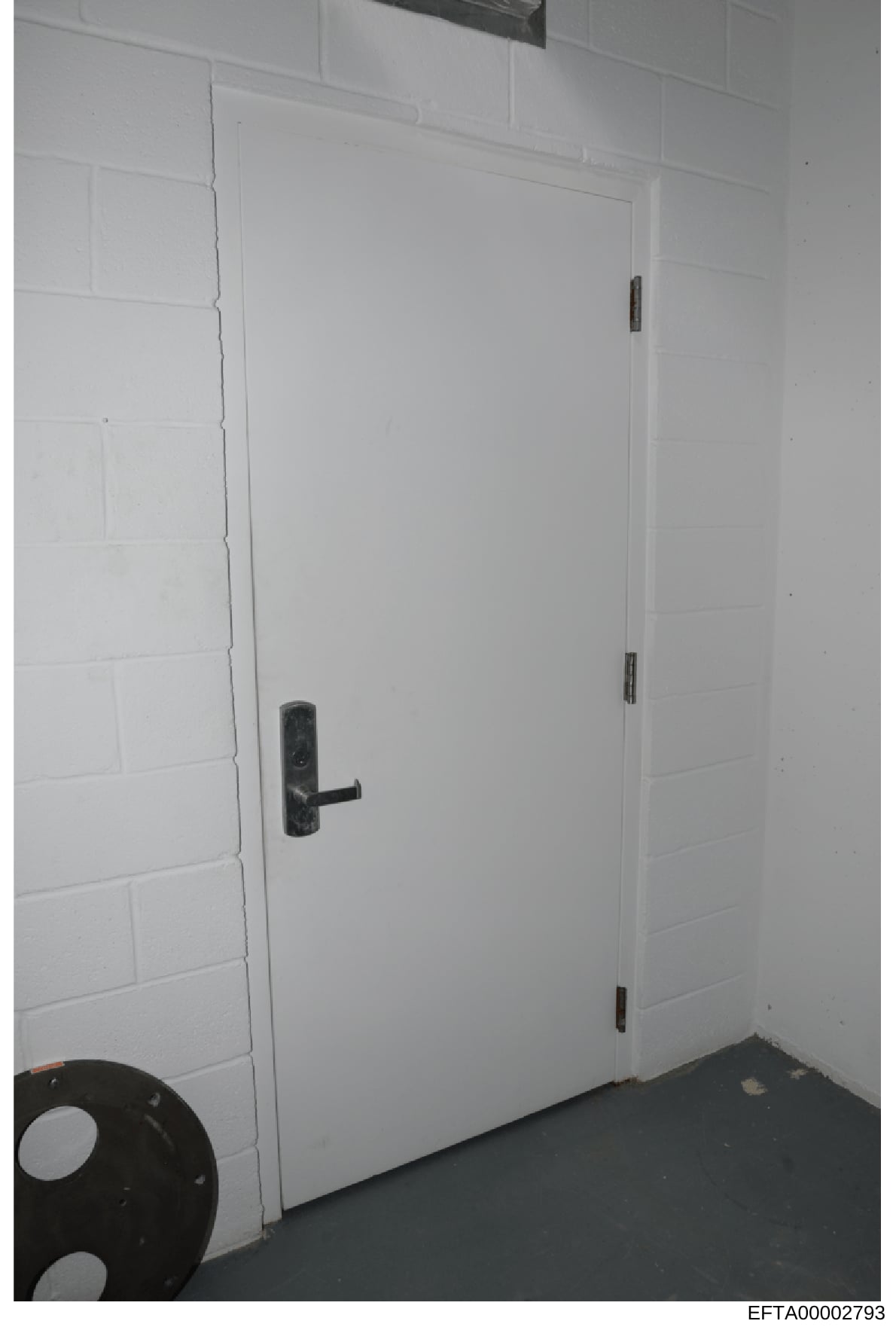 This is an evidence photograph showing the interior of a room with white painted brick walls and a white door with a dark metal handle. A black object (possibly a tire or circular item) is partially visible in the lower left corner. The image appears